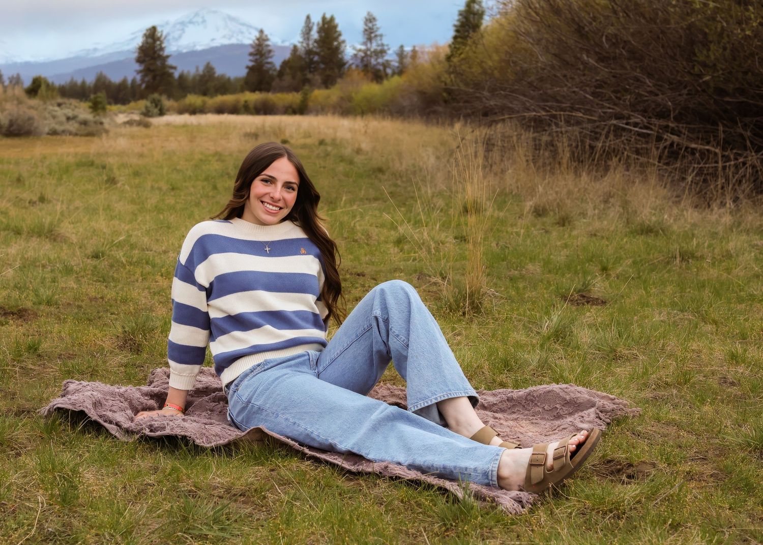Young woman in striped sweater and jeans sitting on blanket in green meadow with snow-capped mountain backdrop.