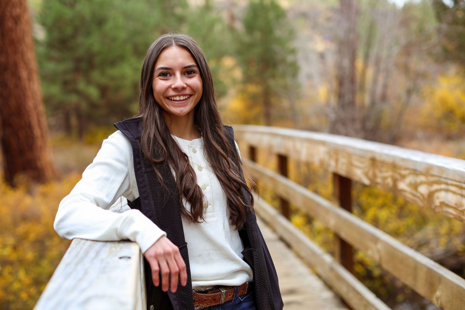 Young woman with long dark hair smiling on a wooden bridge surrounded by autumn foliage in a forest.