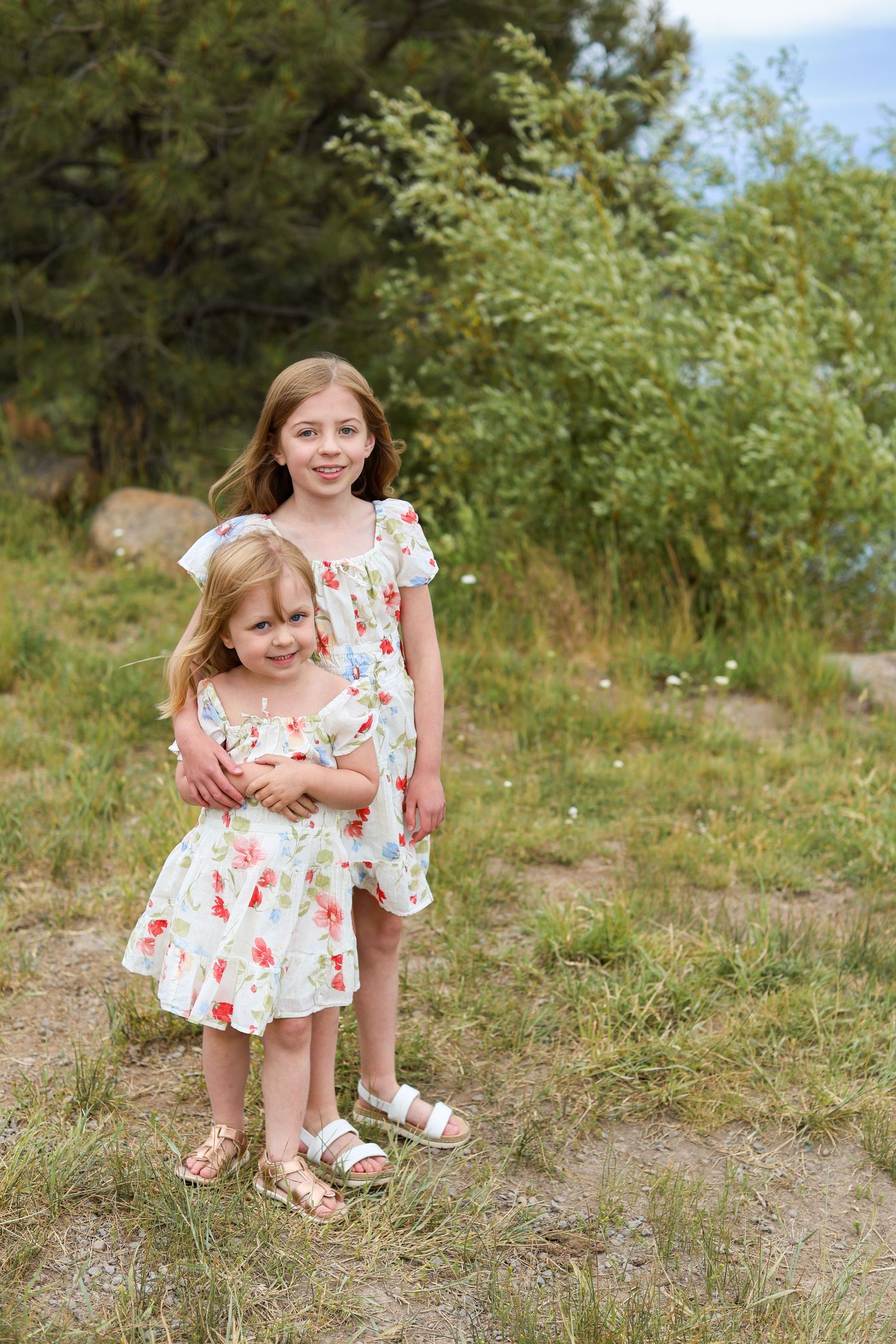 Two young girls in matching floral dresses pose together outdoors on a grassy hillside with green trees behind them. Dillons Falls Bend Or.