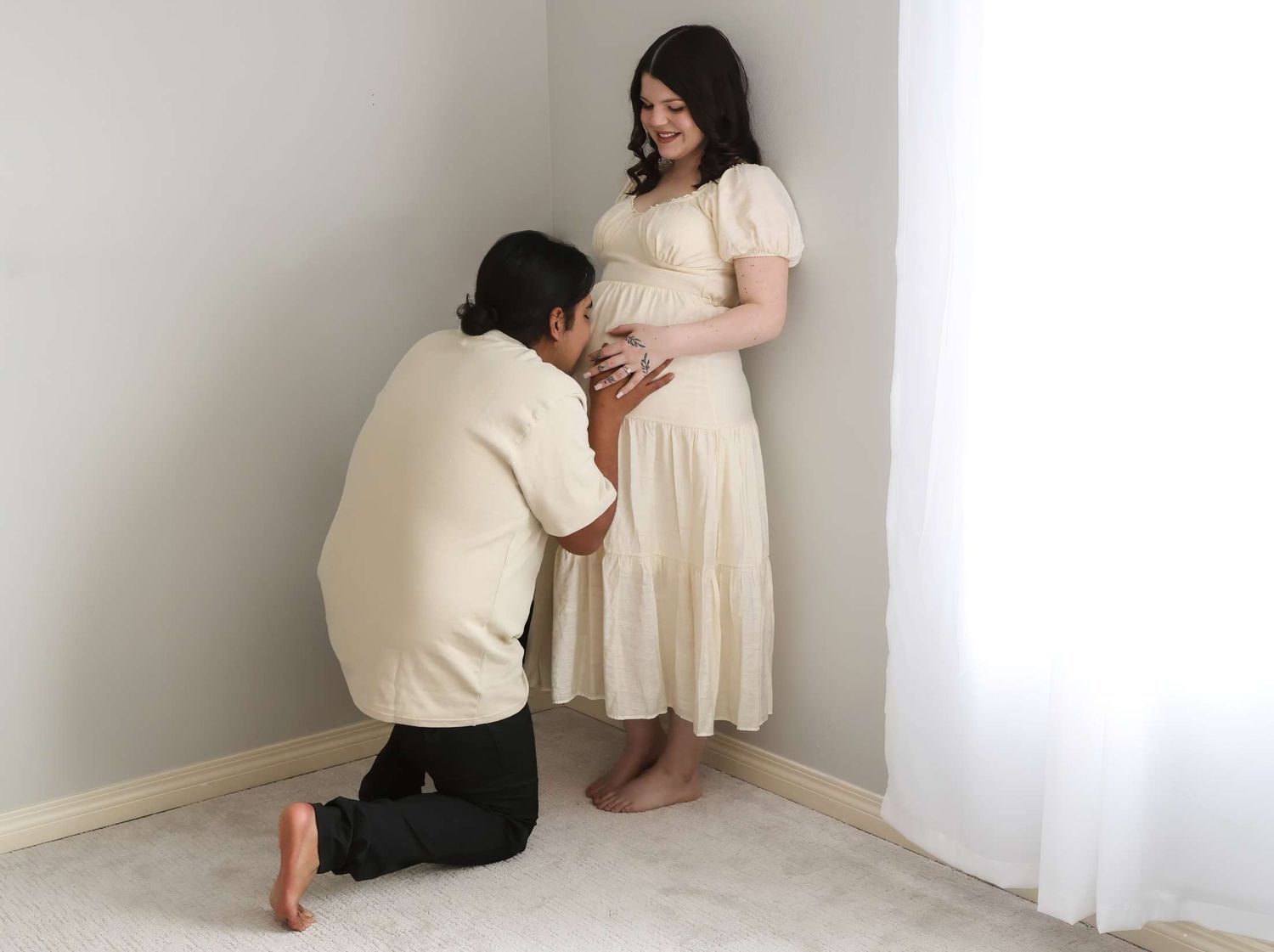 Intimate moment as partner kneels to kiss pregnant belly in white dress.