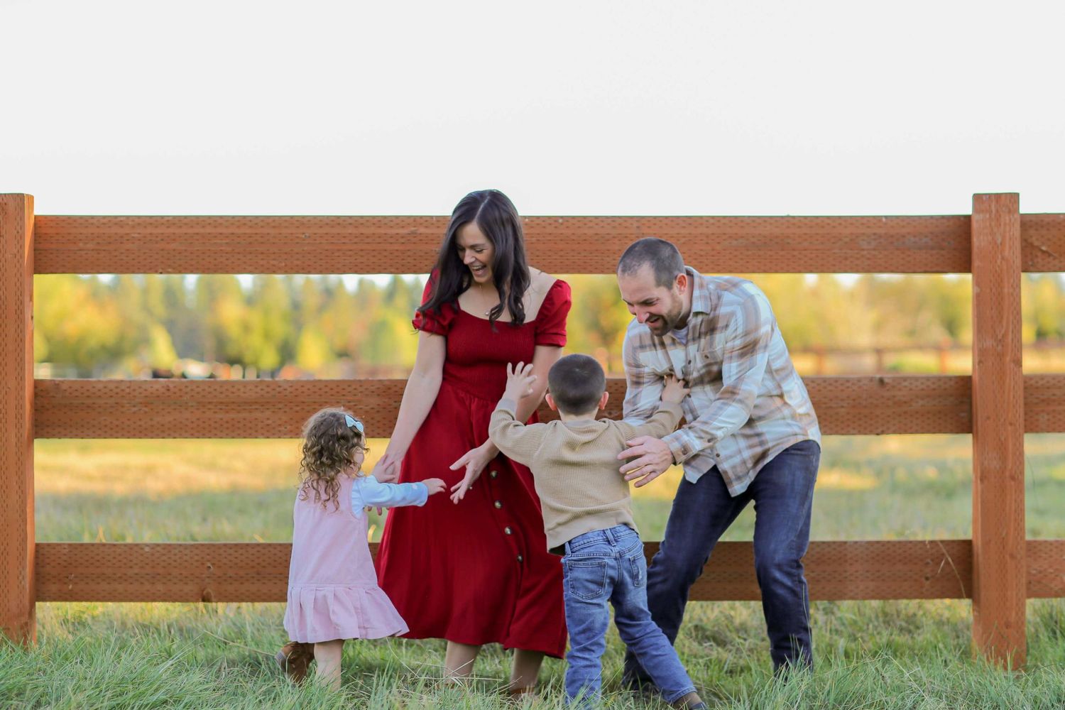 Family of four playing together outdoors near a wooden fence in a green field during golden hour.