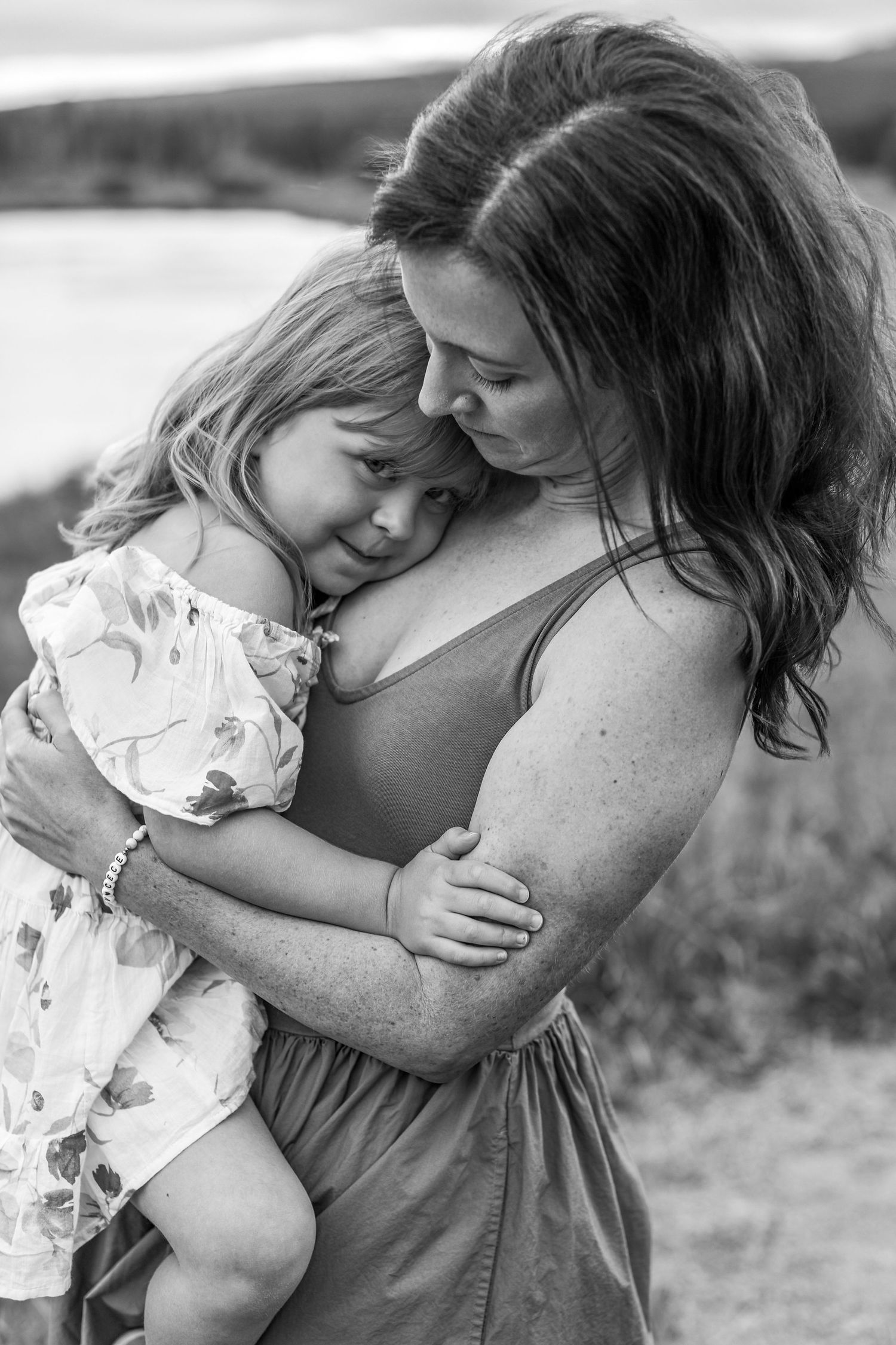 Black and white photo of a mother tenderly embracing her young daughter outdoors near a lake.