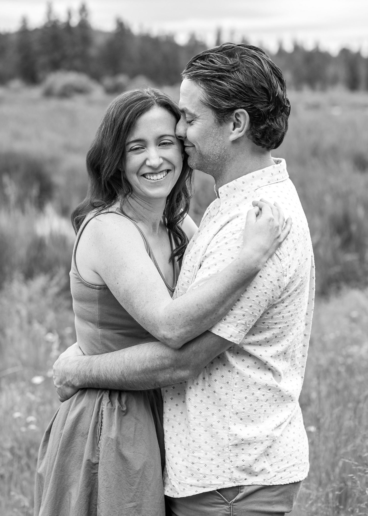 Black and white photo of a smiling couple embracing in a meadow, woman in dress, man in button-up shirt.