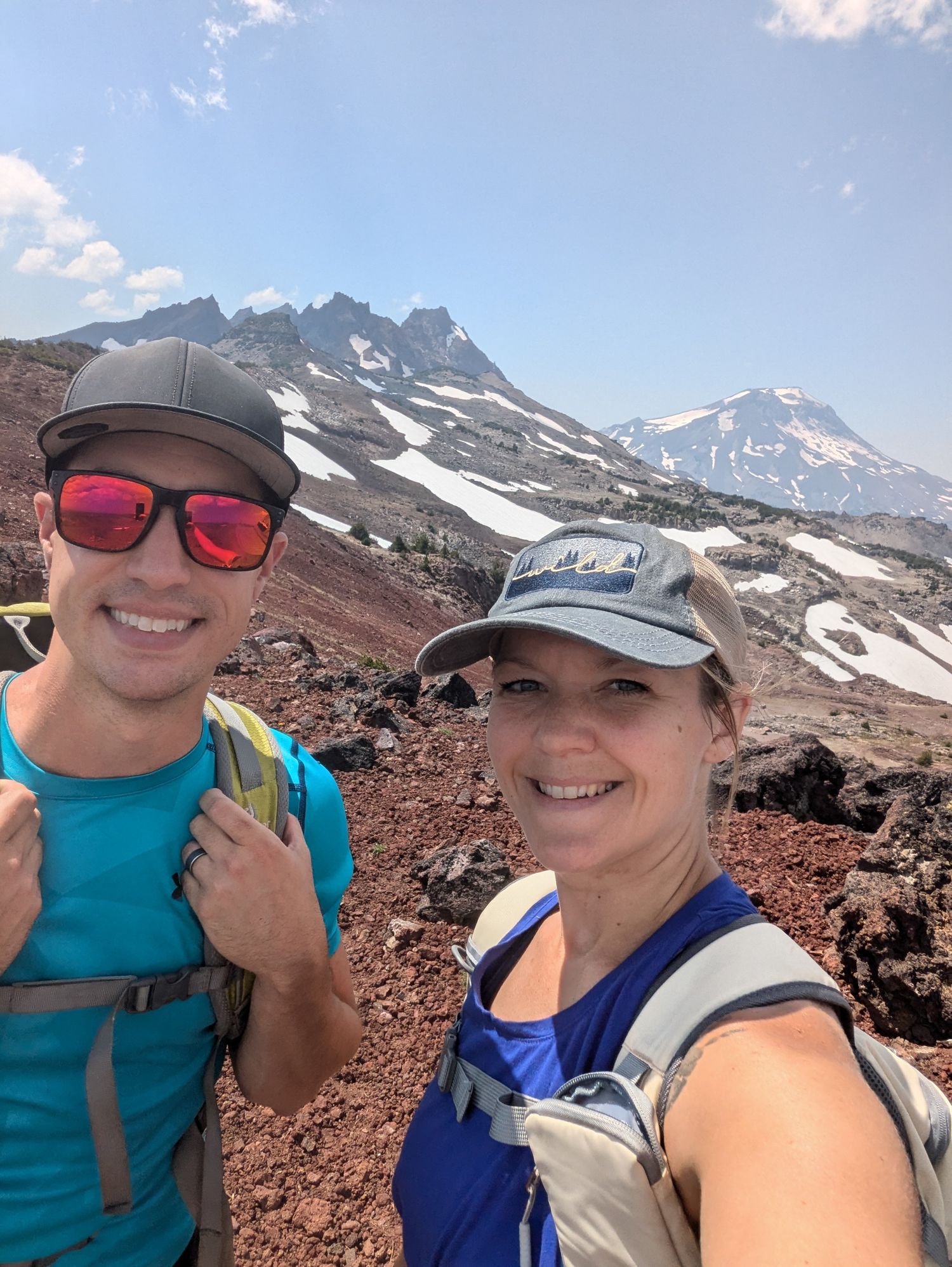 Two hikers smiling for a selfie on a rocky mountain trail with snow-capped peaks and blue sky behind them.
