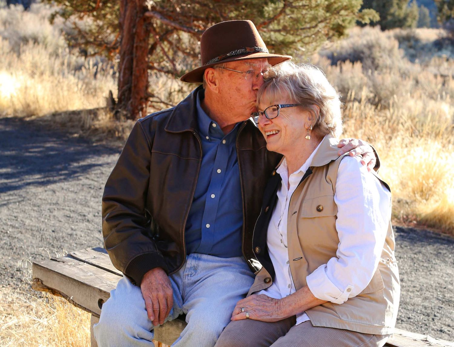 Two people sit together on a wooden bench in a sunny outdoor setting surrounded by dry grass and trees.