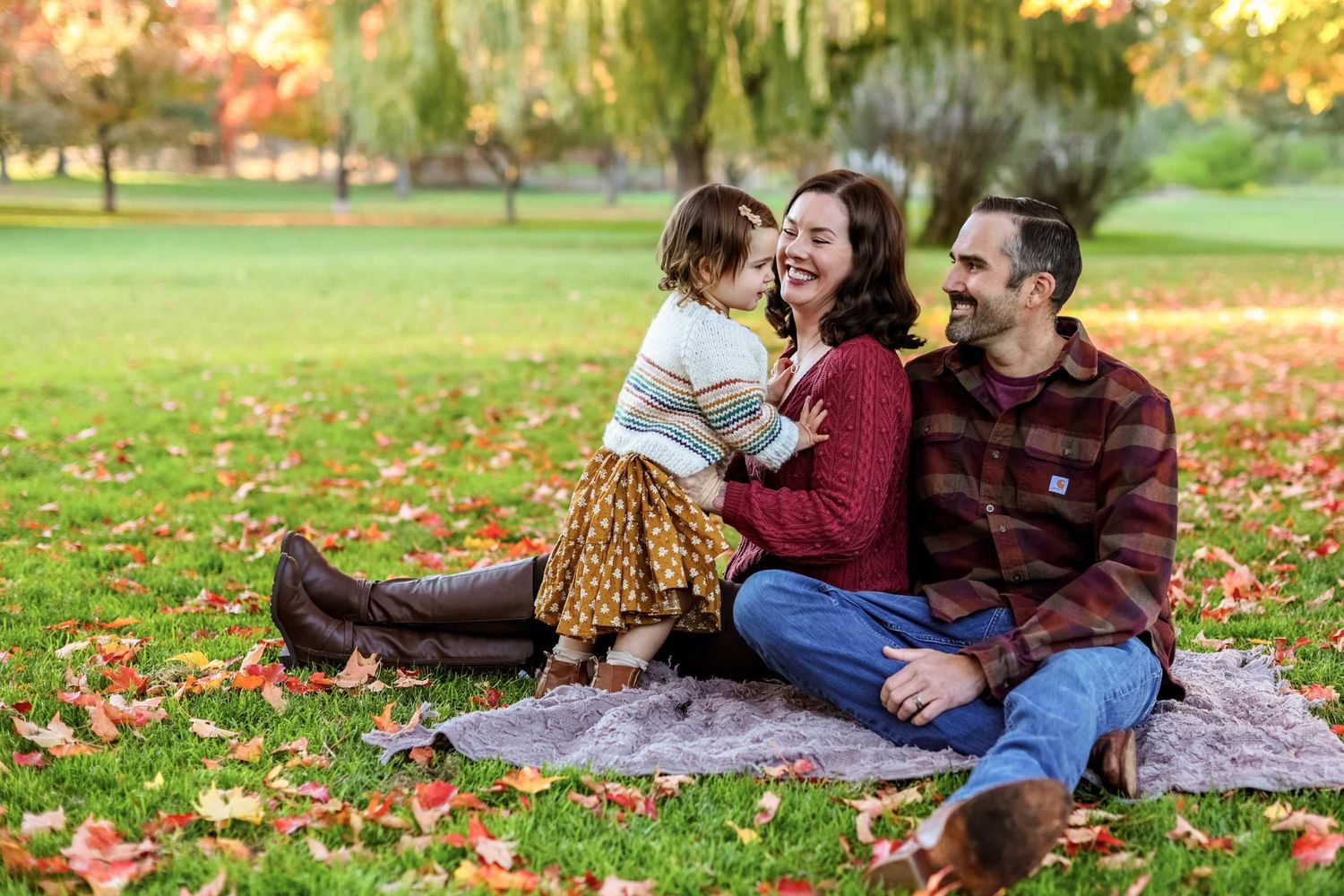 Couple sits together on plaid blanket surrounded by colorful autumn leaves in a peaceful park setting.