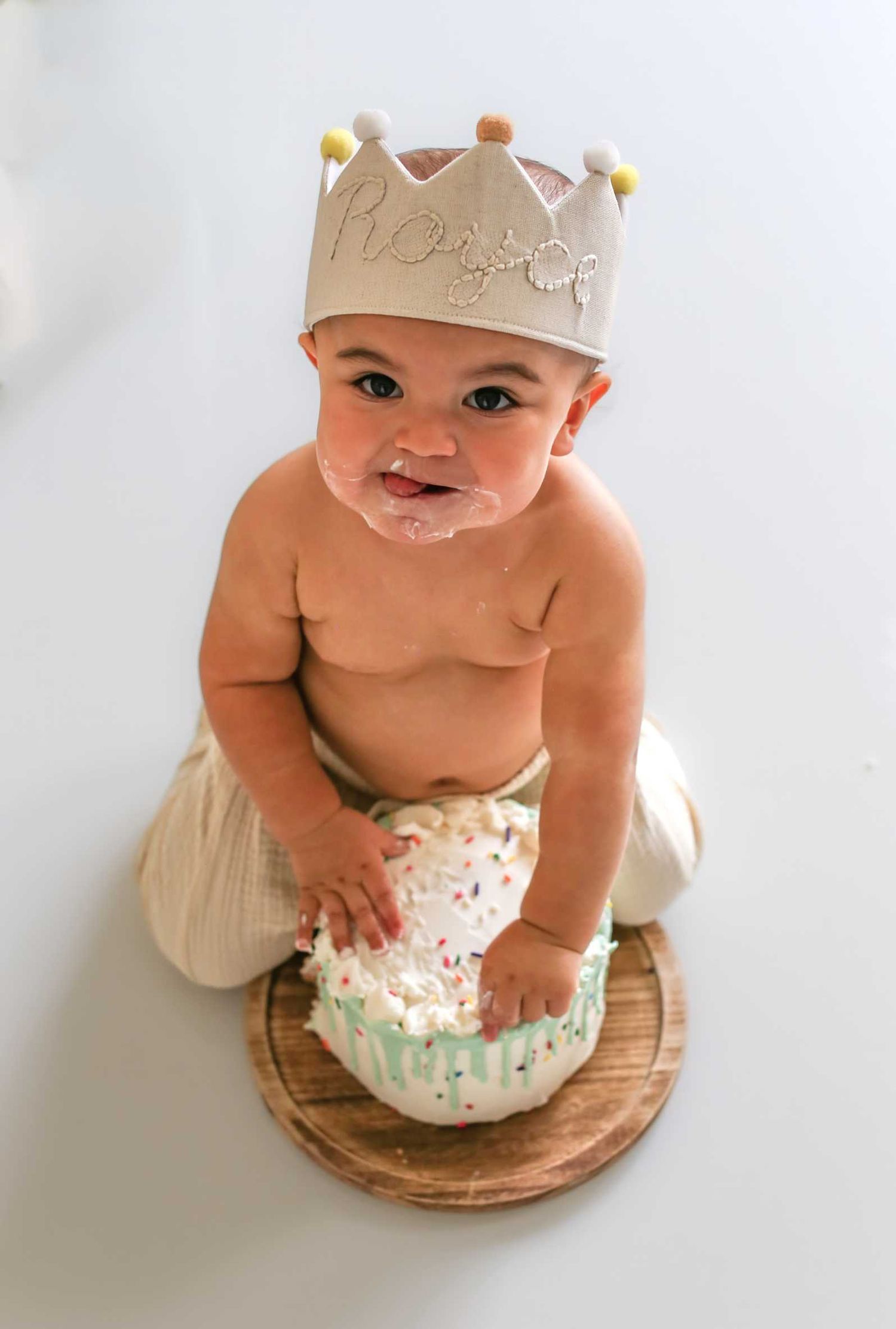Baby wearing crown sits on wooden platform during milestone photoshoot.