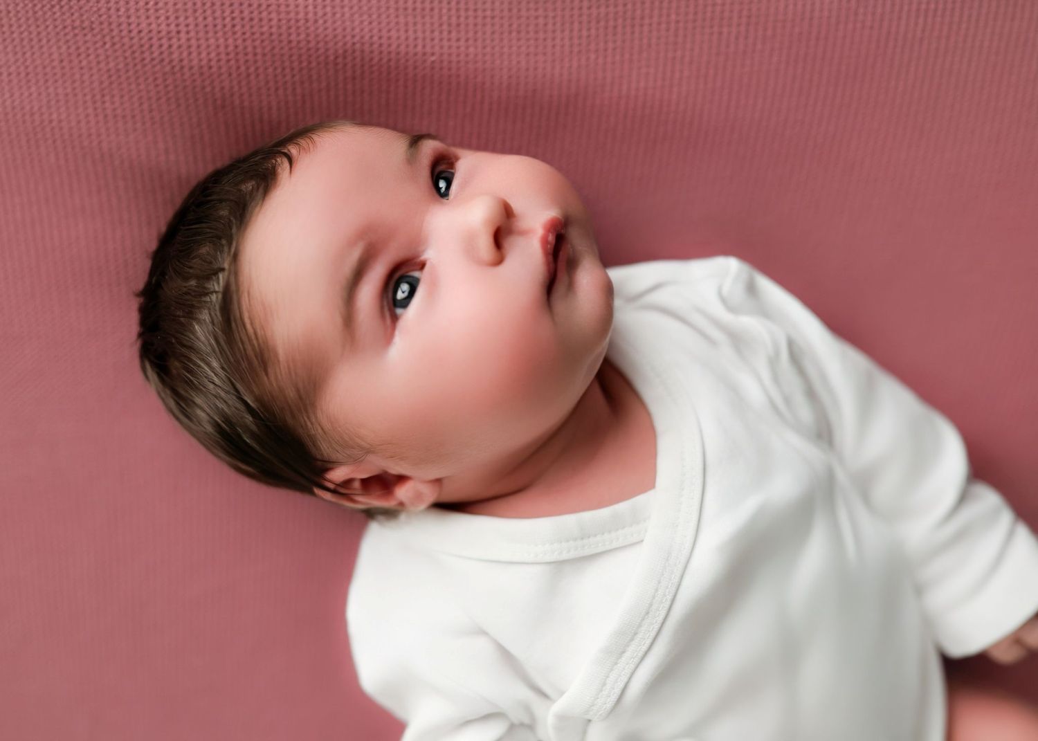 Baby in white onesie lying on pink background looking up with curious expression.