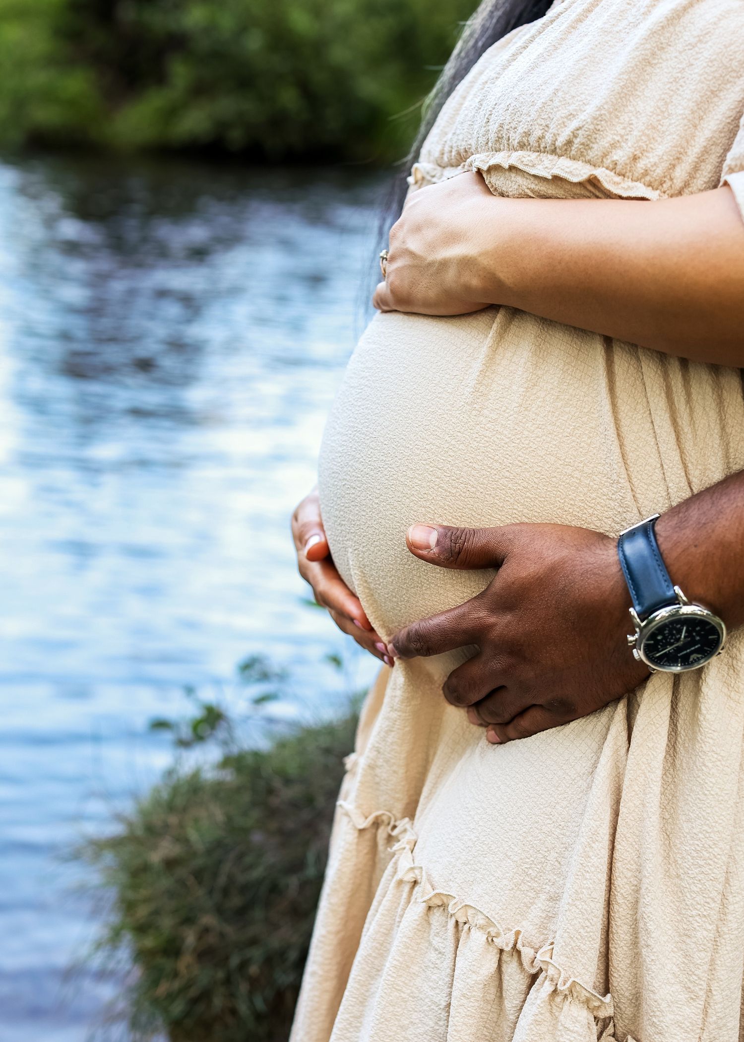 Pregnant woman in beige dress cradling baby bump outdoors near a river, with partner's hand wearing a blue watch.