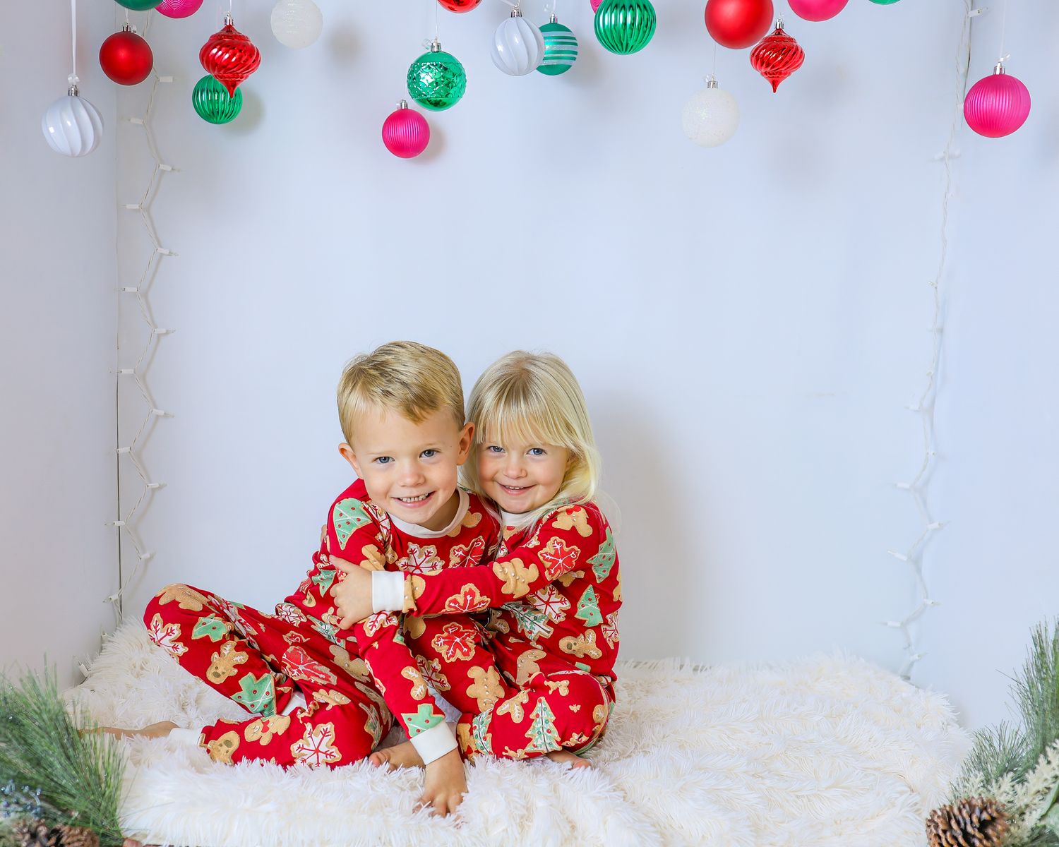 Two smiling children in matching red holiday pajamas hug on a fluffy white blanket beneath hanging Christmas ornaments.
