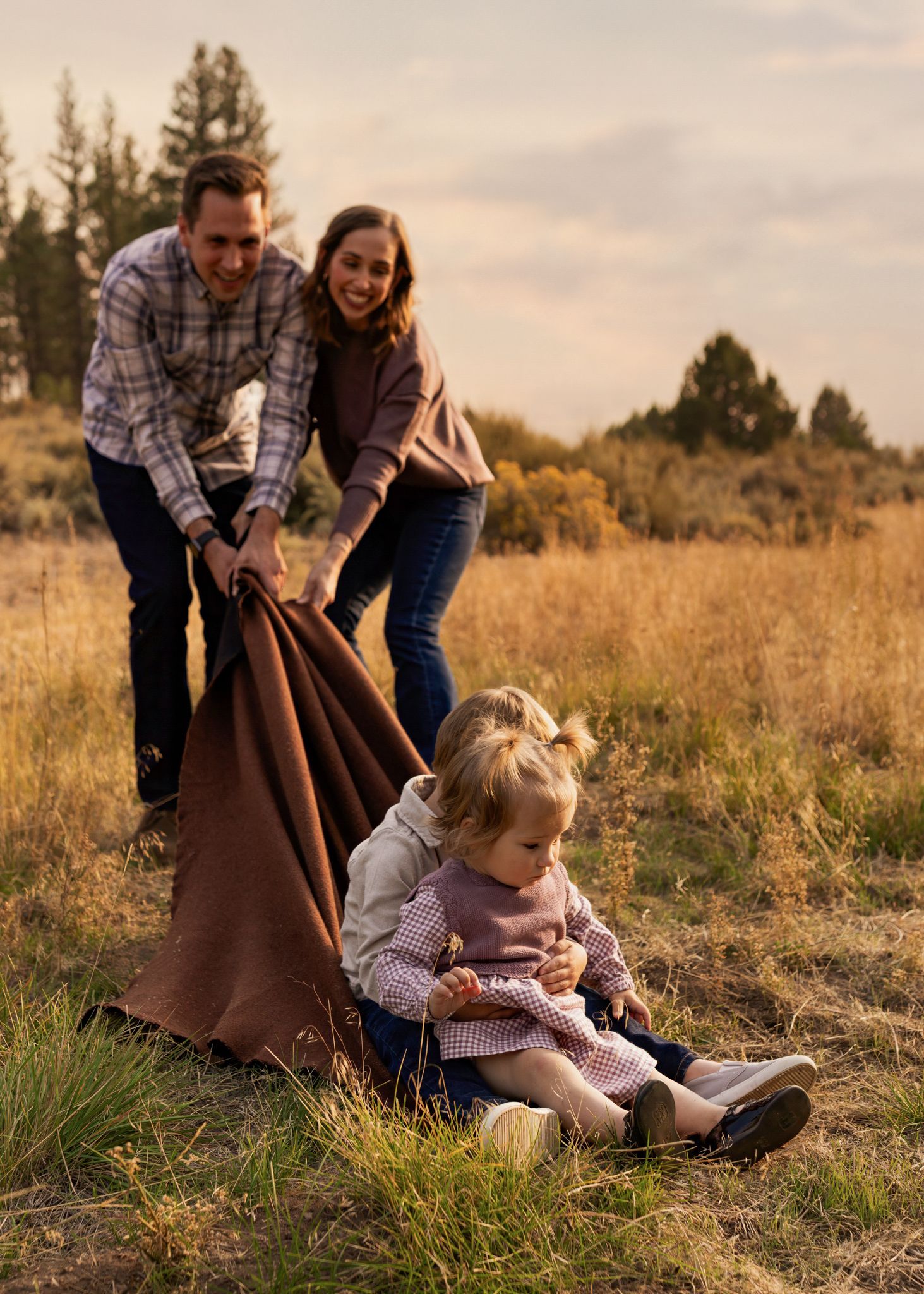 A family laughs while pulling a brown blanket with a toddler sitting on it in a golden field.