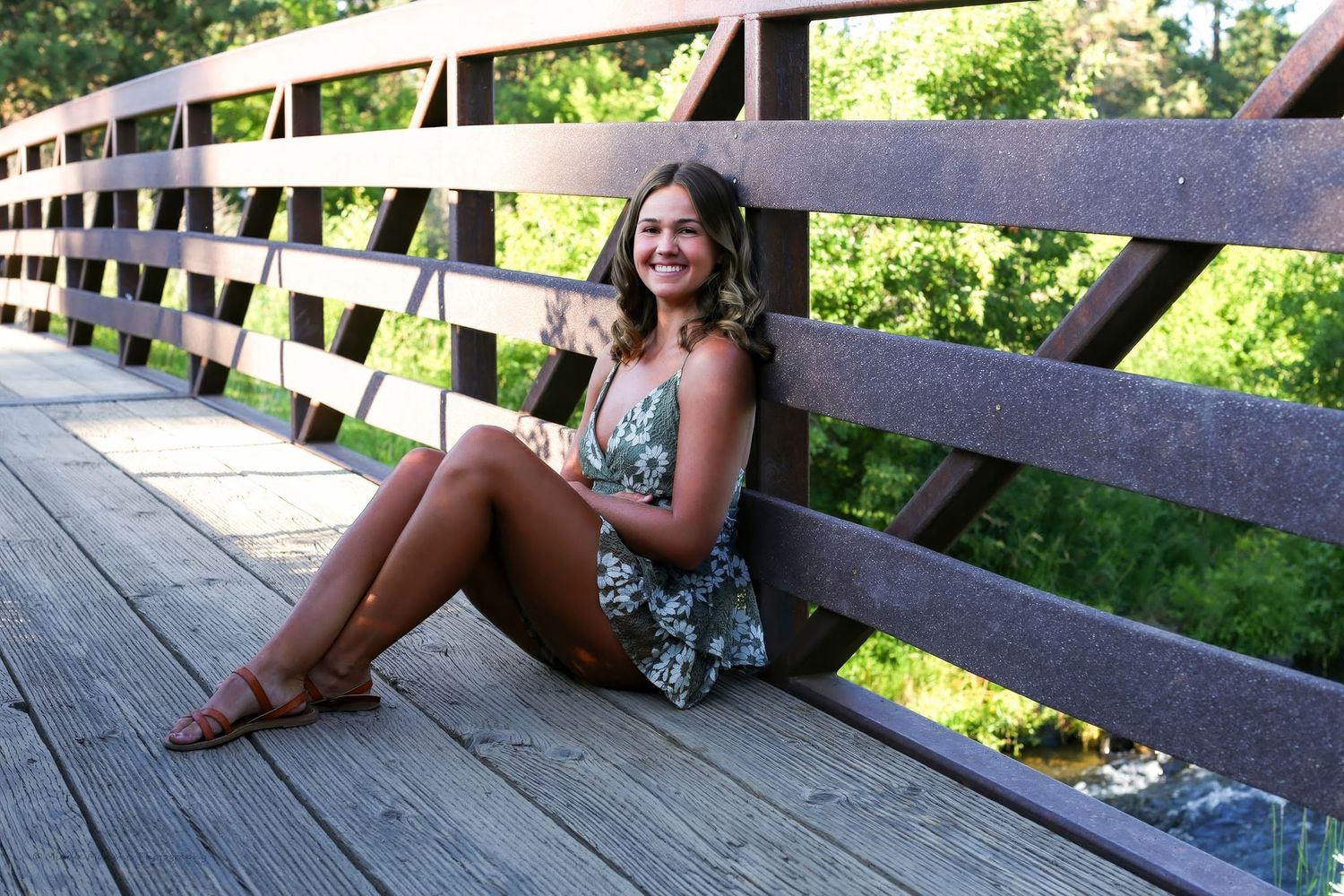 Young woman in floral romper sitting on wooden bridge, smiling outdoors surrounded by lush green trees.