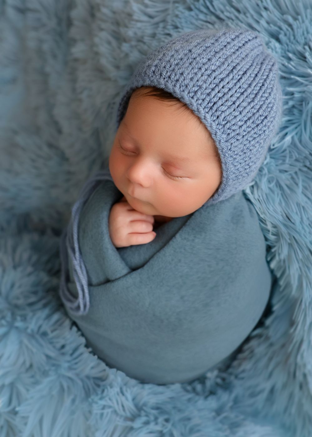 Sleeping newborn baby swaddled in blue wrap wearing knit bonnet hat on fluffy blue backdrop.