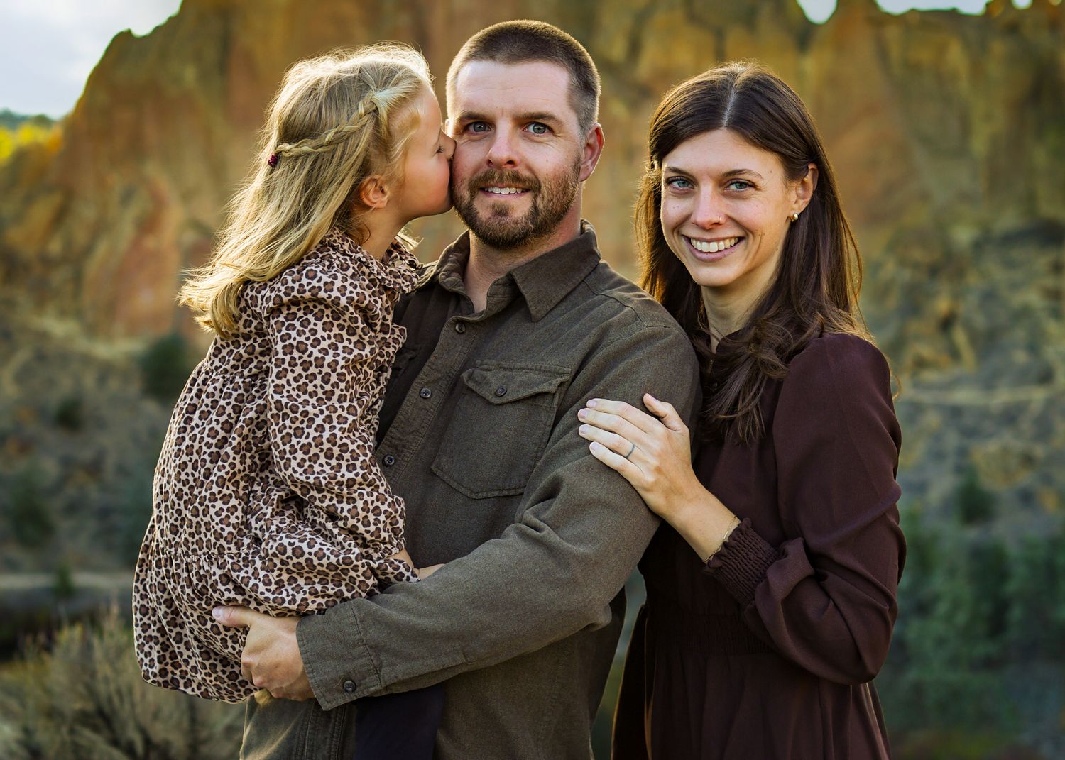 A happy family of three posing outdoors, with a young girl kissing her father on the cheek while mother smiles.
