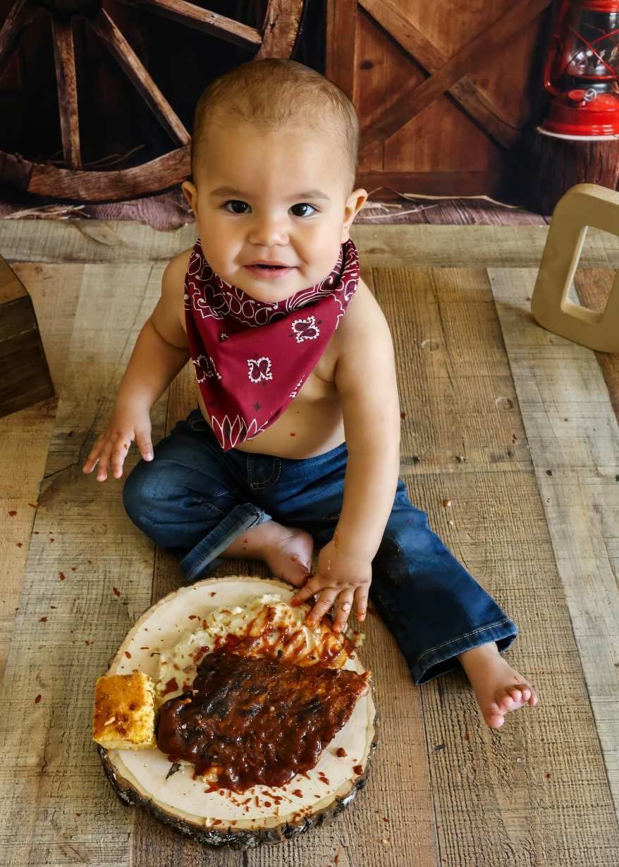 Cute baby in red bandana and jeans sitting on wood floor with BBQ ribs plate at cowboy themed first birthday. Redmond Oregon Studio