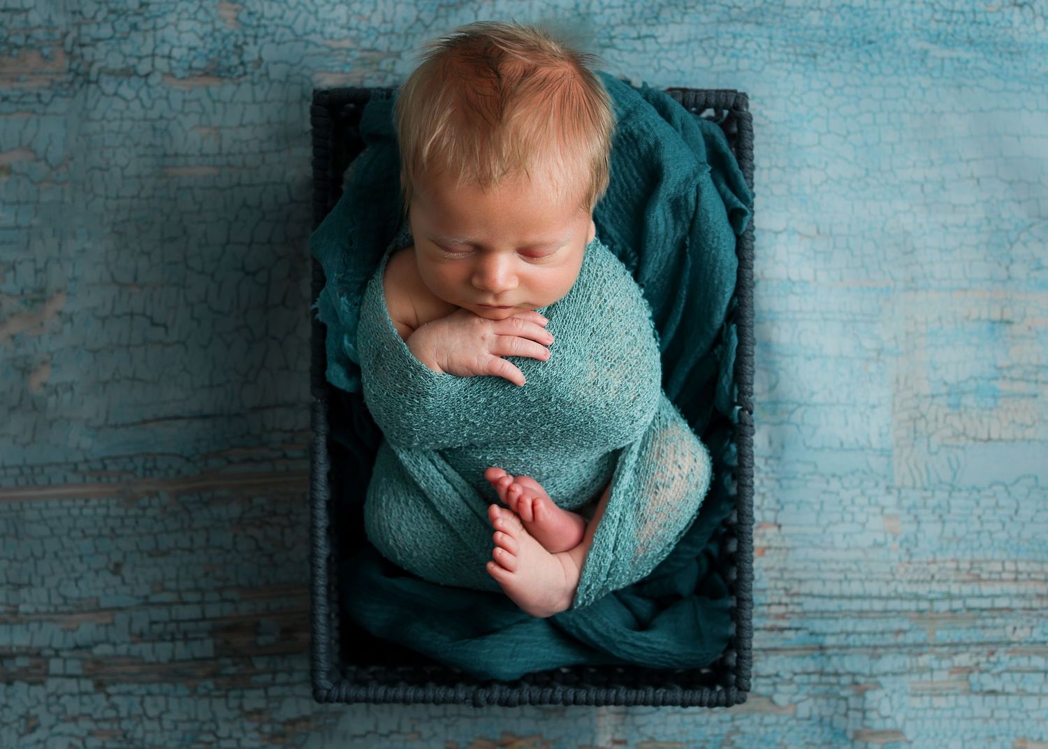 Newborn baby swaddled in teal wrap sleeping peacefully in a dark wooden box on a turquoise background.