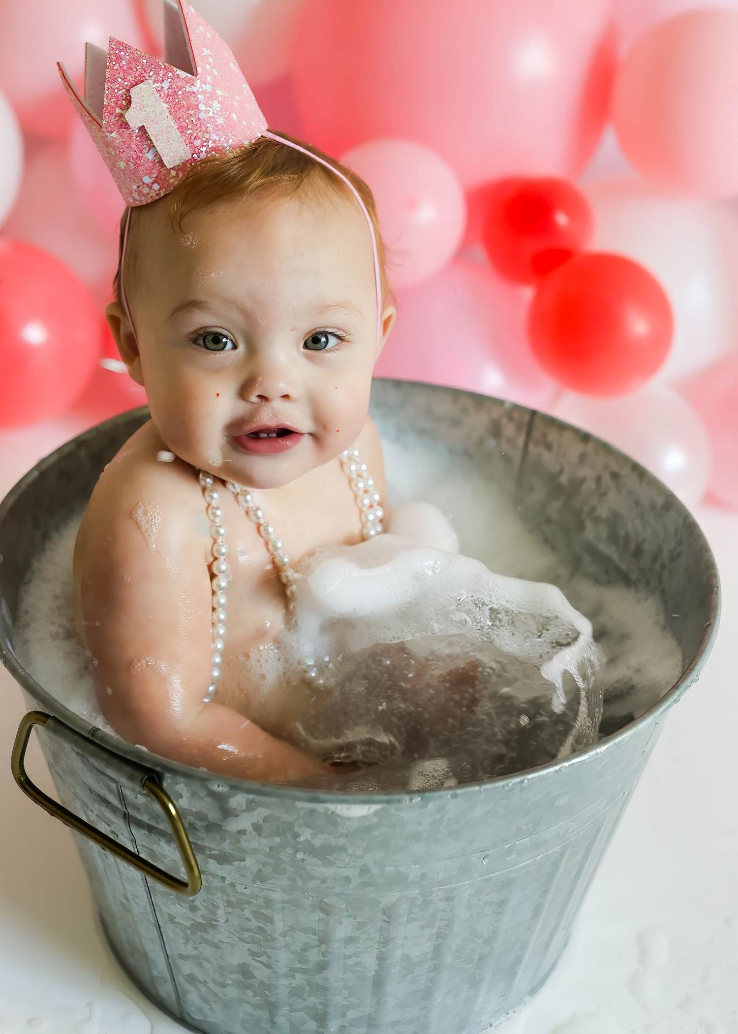 Baby bath time photo with pink crown and bubbles in galvanized metal tub with pink balloon backdrop.