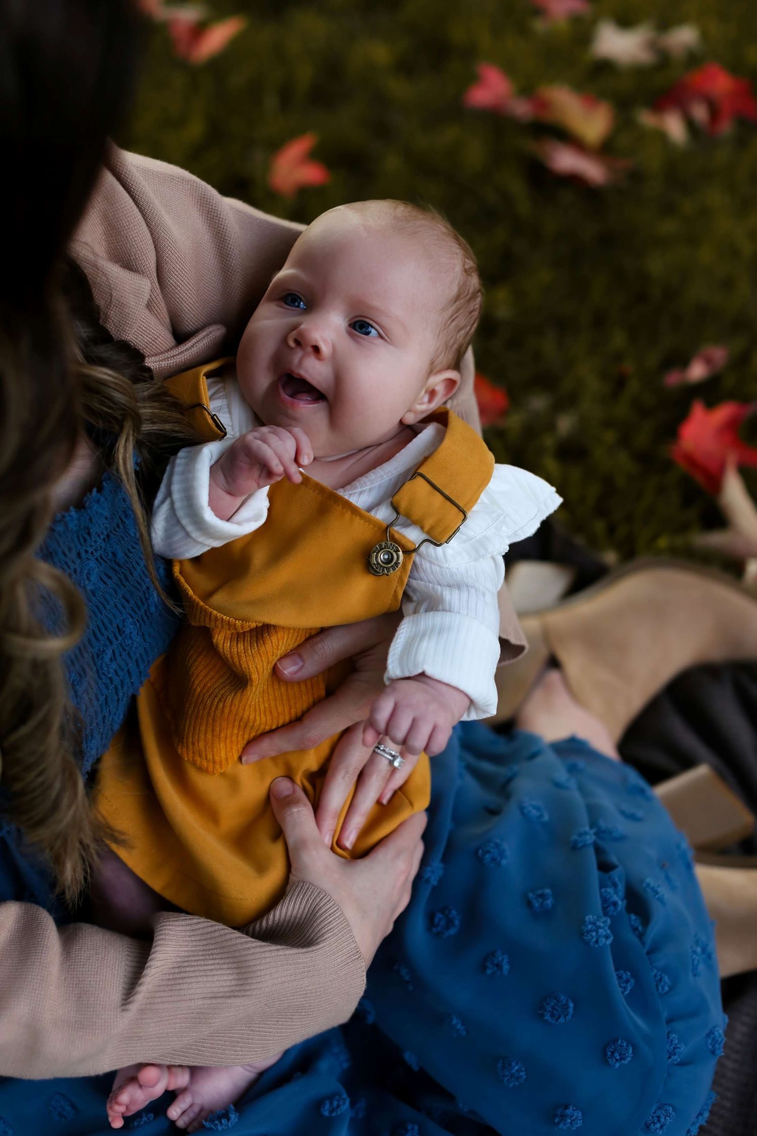 Baby wearing yellow sweater and blue dress plays with a toy while laying on autumn leaves.