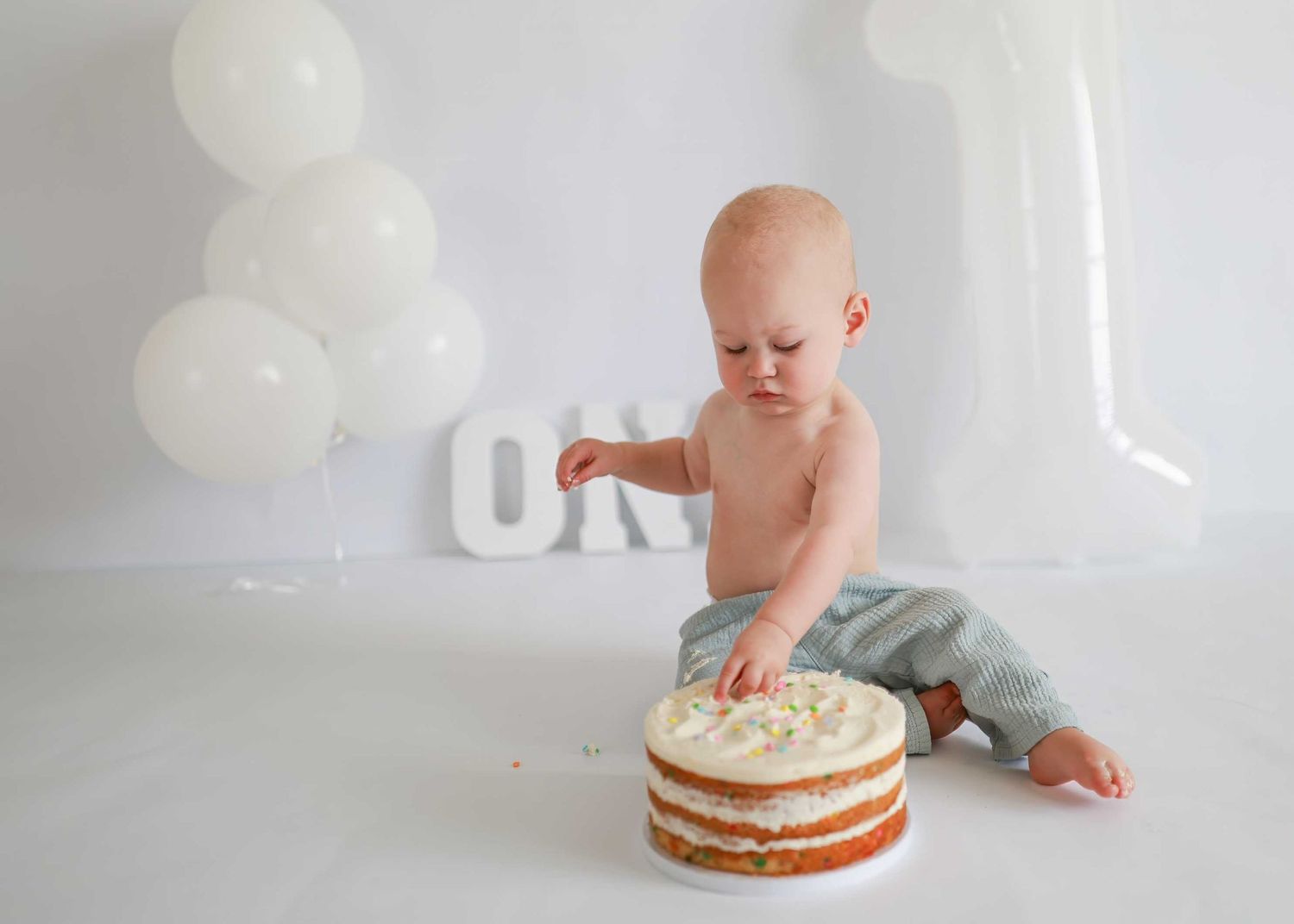 Baby in jeans exploring a white frosted birthday cake with white balloons in the background.