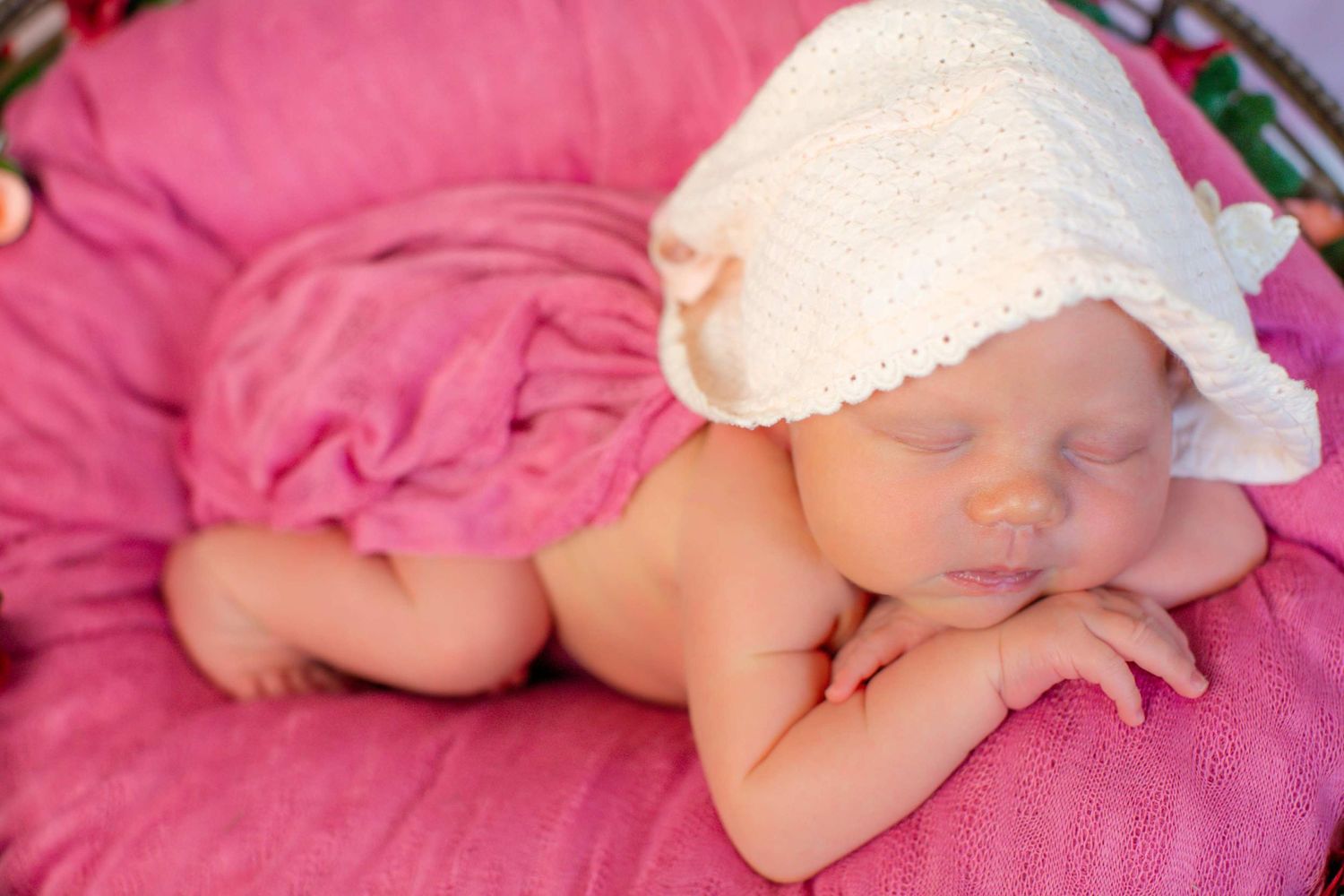 Sleeping newborn in white bonnet resting on vibrant pink fabric.