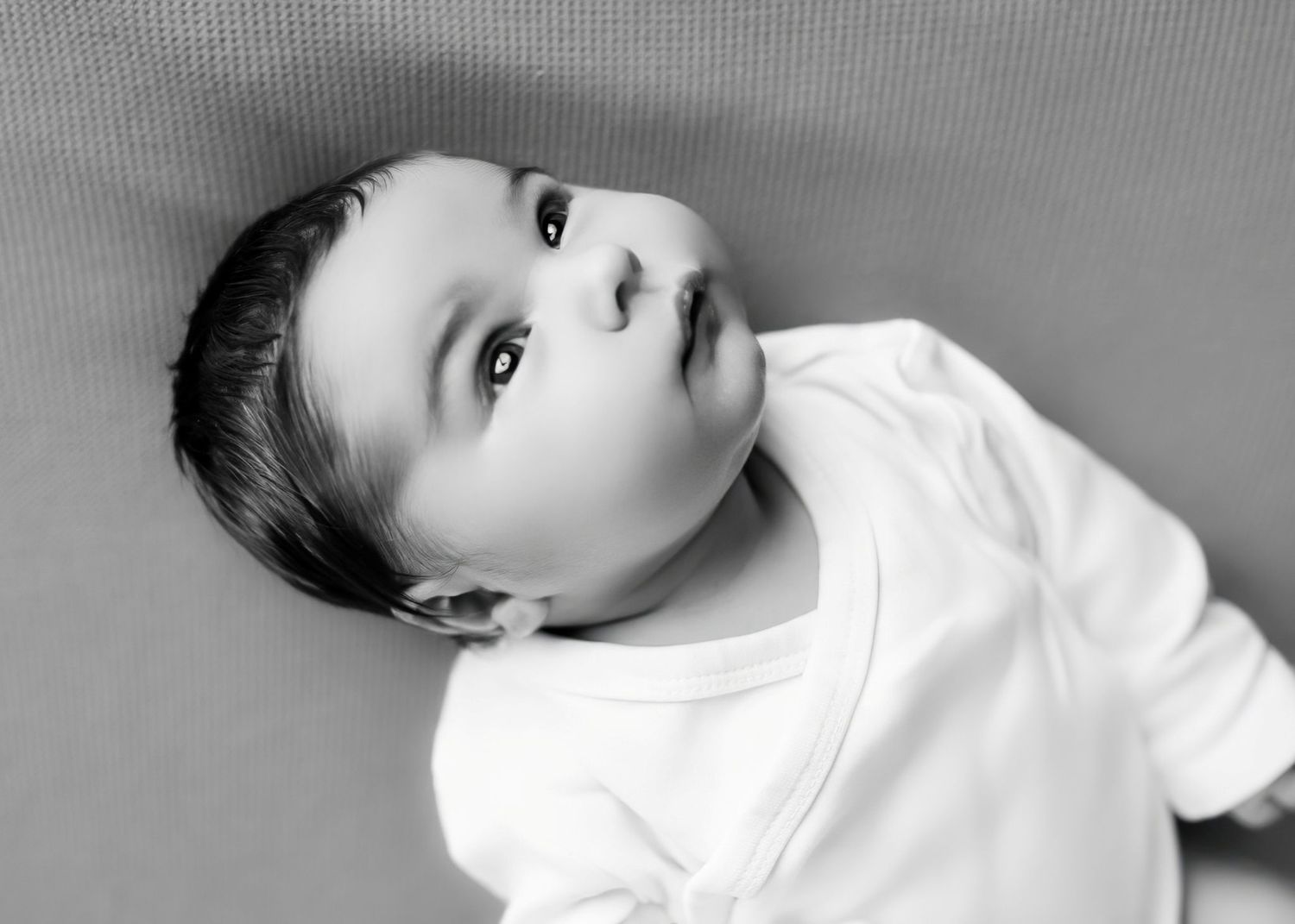 Black and white photo of an adorable baby in a white onesie looking up with big eyes. Redmond Oregon Studio