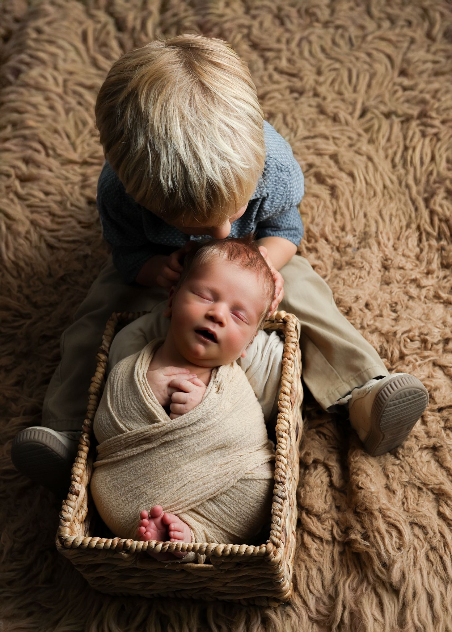 A young blonde boy gently kisses his sleeping newborn sibling wrapped in cream swaddle inside a wicker basket.