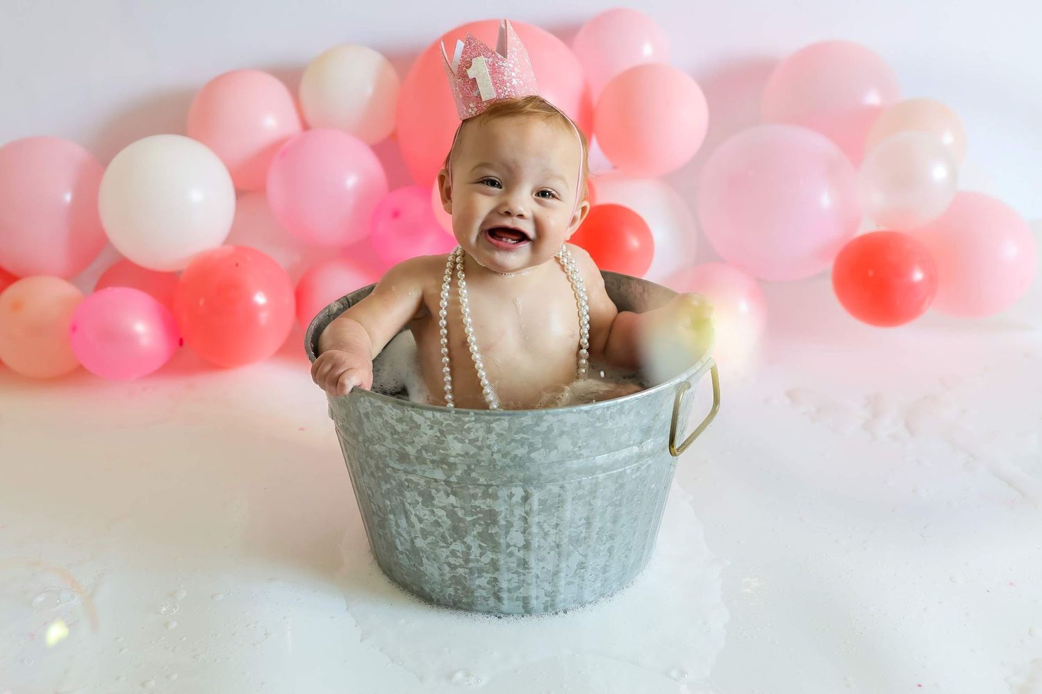 Baby sitting in a grey basket with pink and white balloons in the background during a photo session.