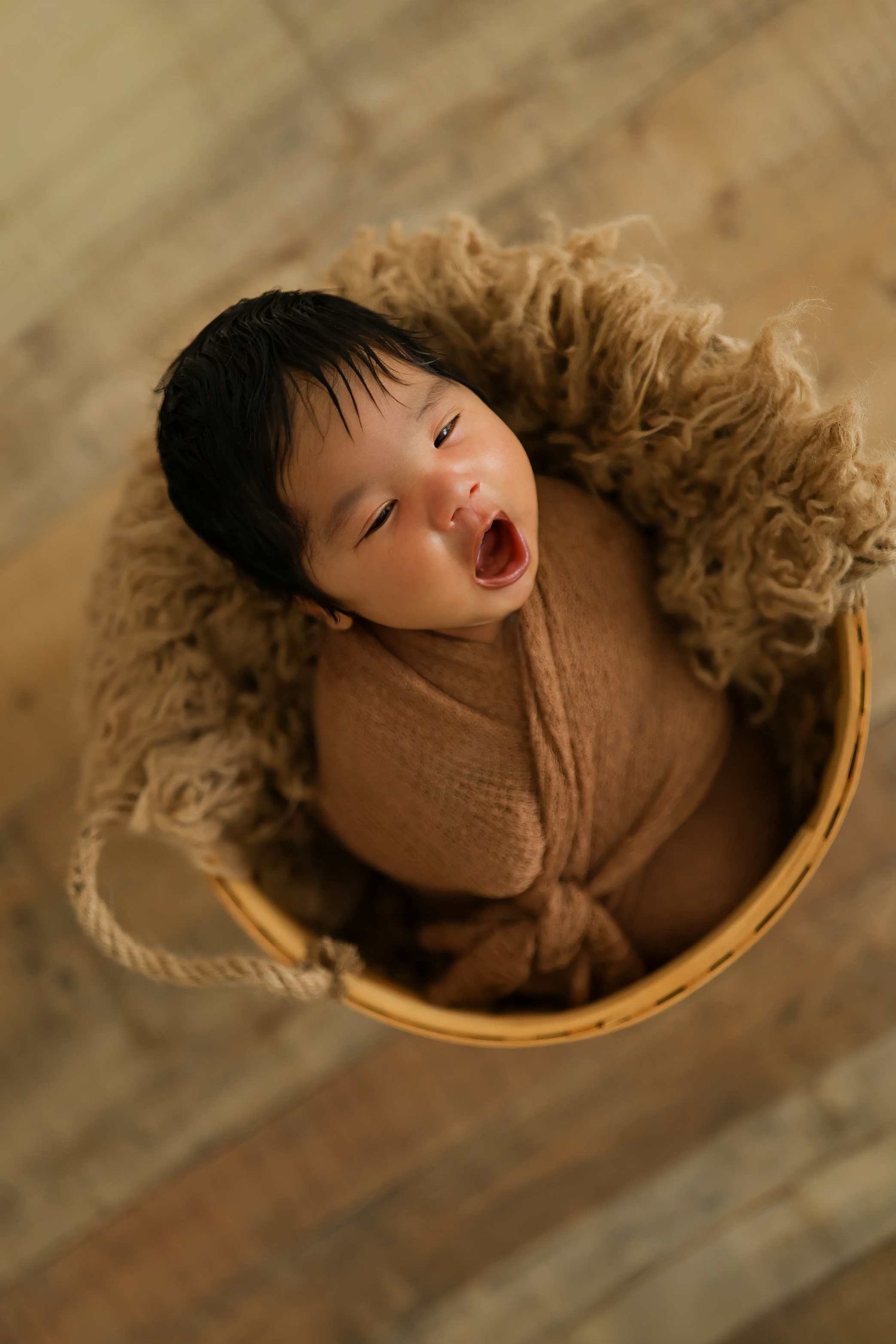 Newborn baby wrapped in brown swaddle, yawning in a rustic wicker basket lined with fluffy tan wool.
