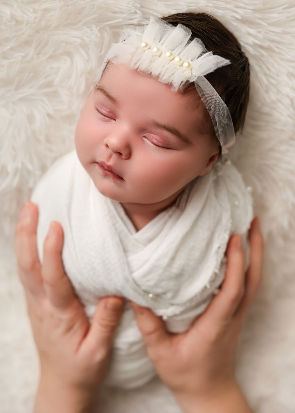 Sleeping newborn baby girl wrapped in white swaddle with pearl headband, cradled in adult hands on fluffy white backdrop.