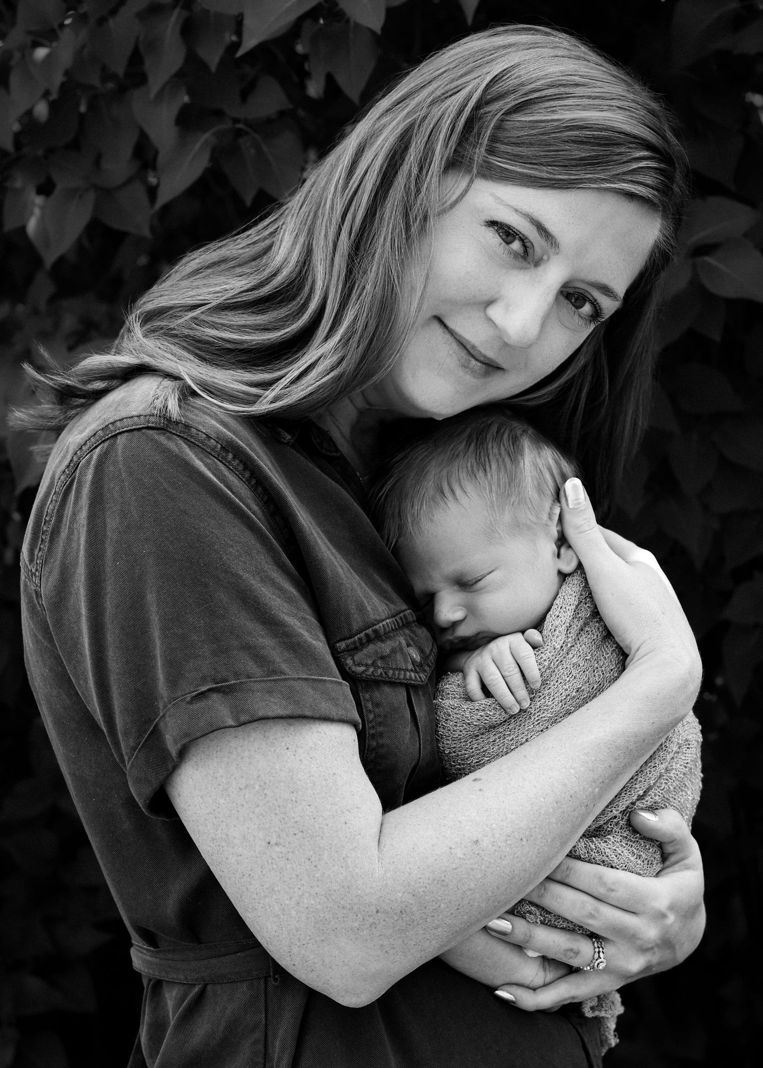 Black and white photo of a smiling woman tenderly cradling a sleeping newborn baby outdoors near lush foliage.