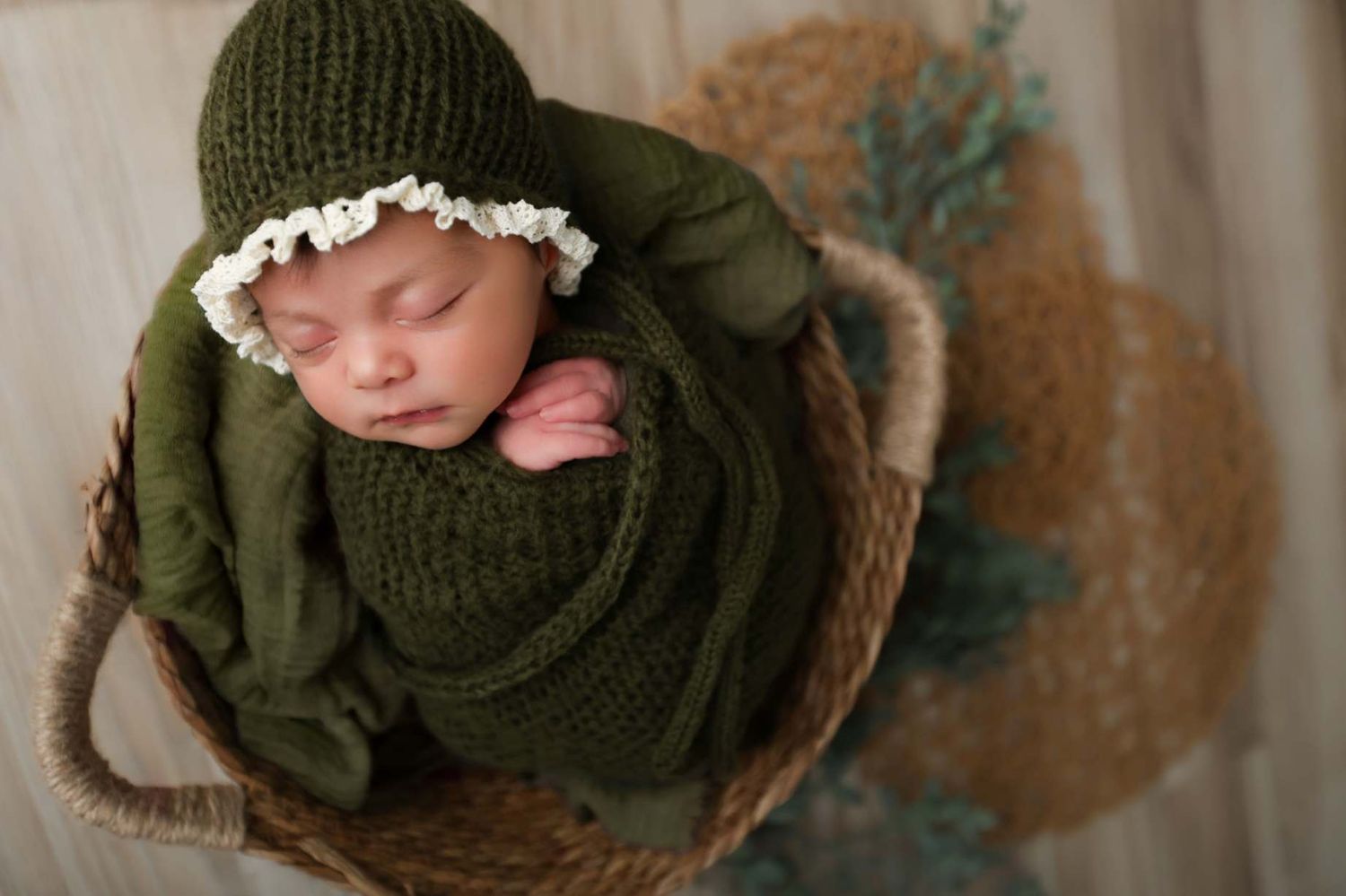 Newborn baby wrapped in green knit blanket and bonnet sleeping peacefully in a wicker basket.