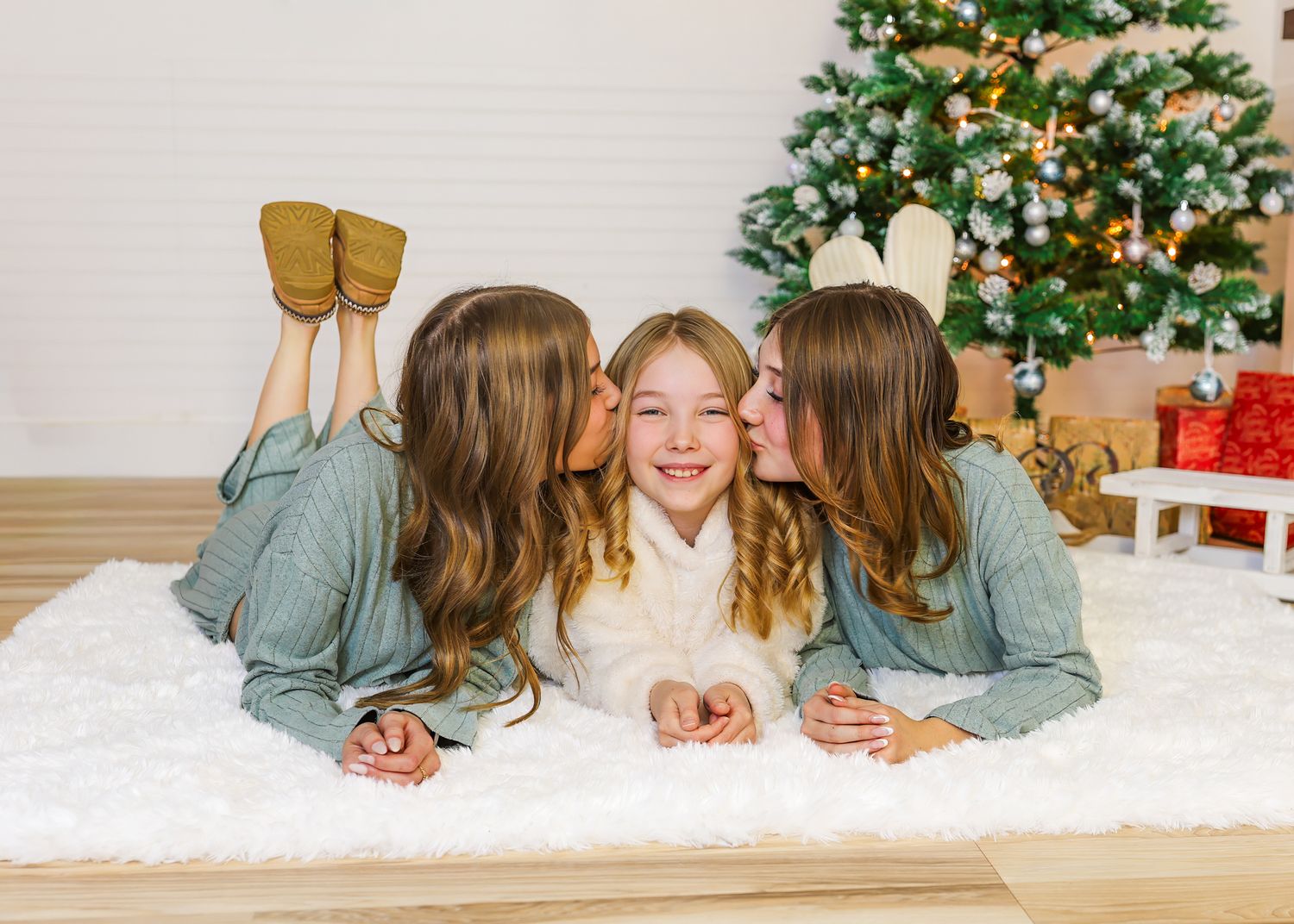 Two girls kiss the cheeks of a smiling girl between them as they lie on a white fur rug in front of a decorated Christmas tree.