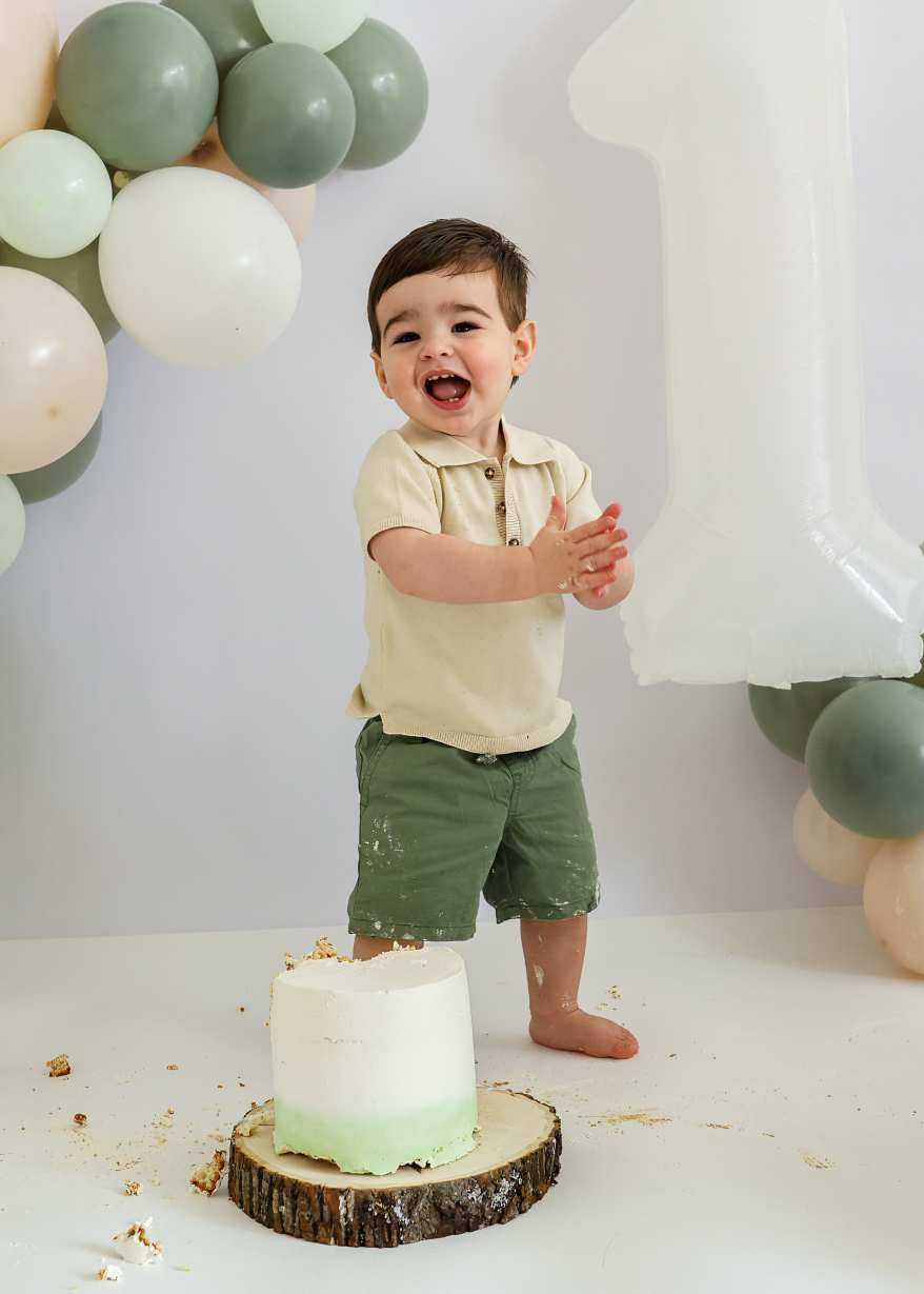 A festive cake smash photo with sage green and white balloons on a rustic wood slice cake stand.