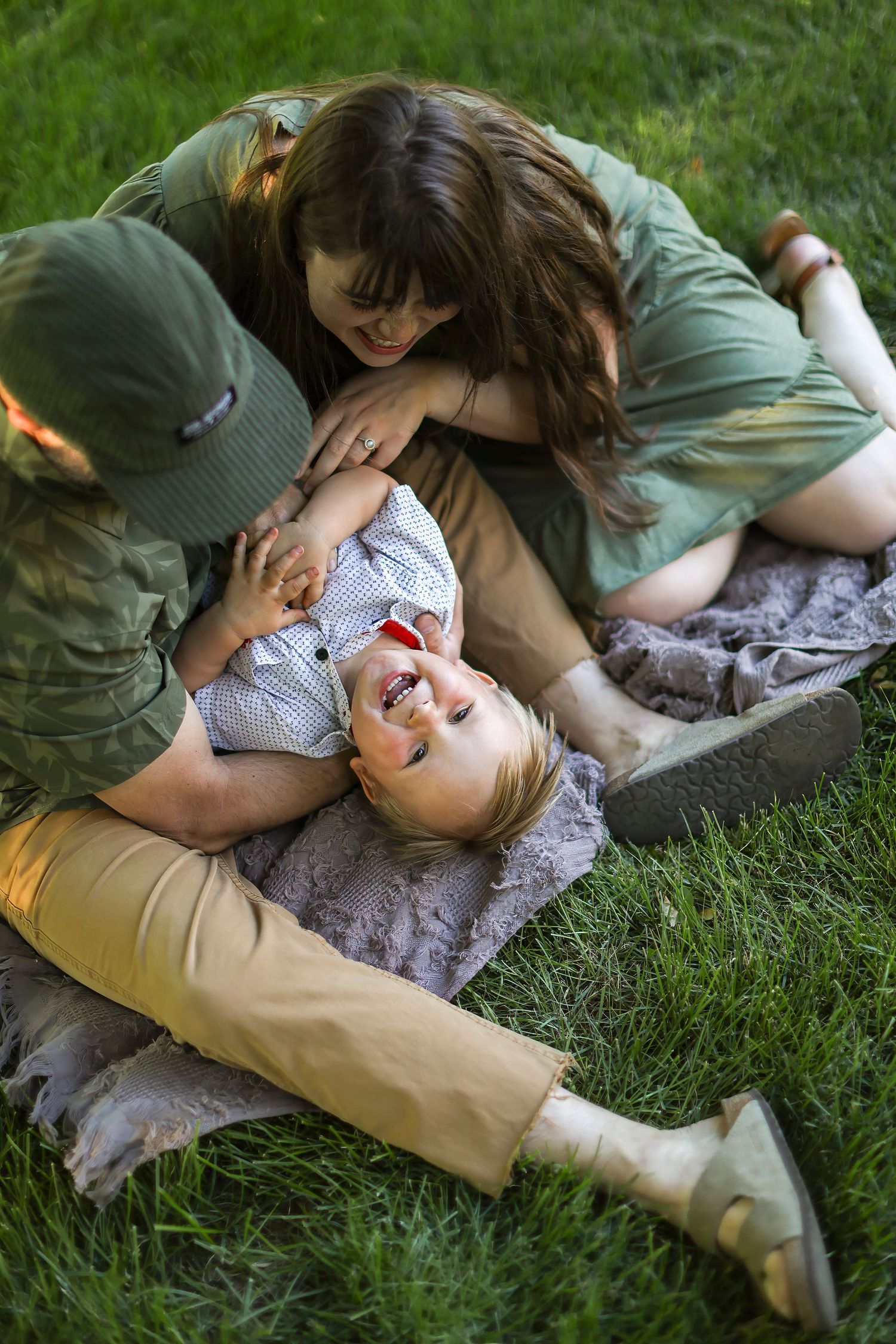 Family laughing and playing together on green grass, parents cuddling joyful toddler on a blanket outdoors.