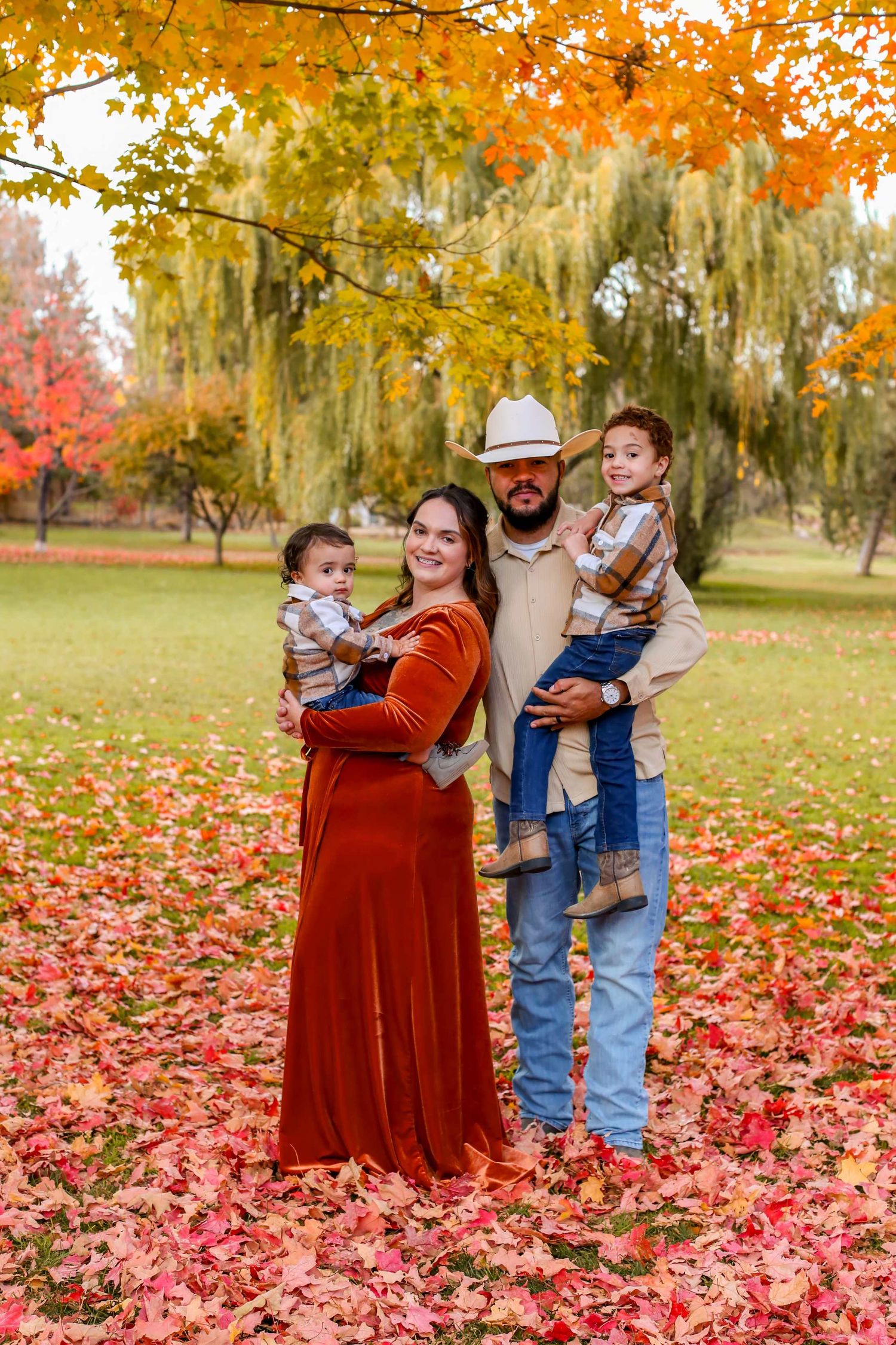 Family of four posing outdoors in fall foliage, parents holding two young children among vibrant red and orange autumn leaves.