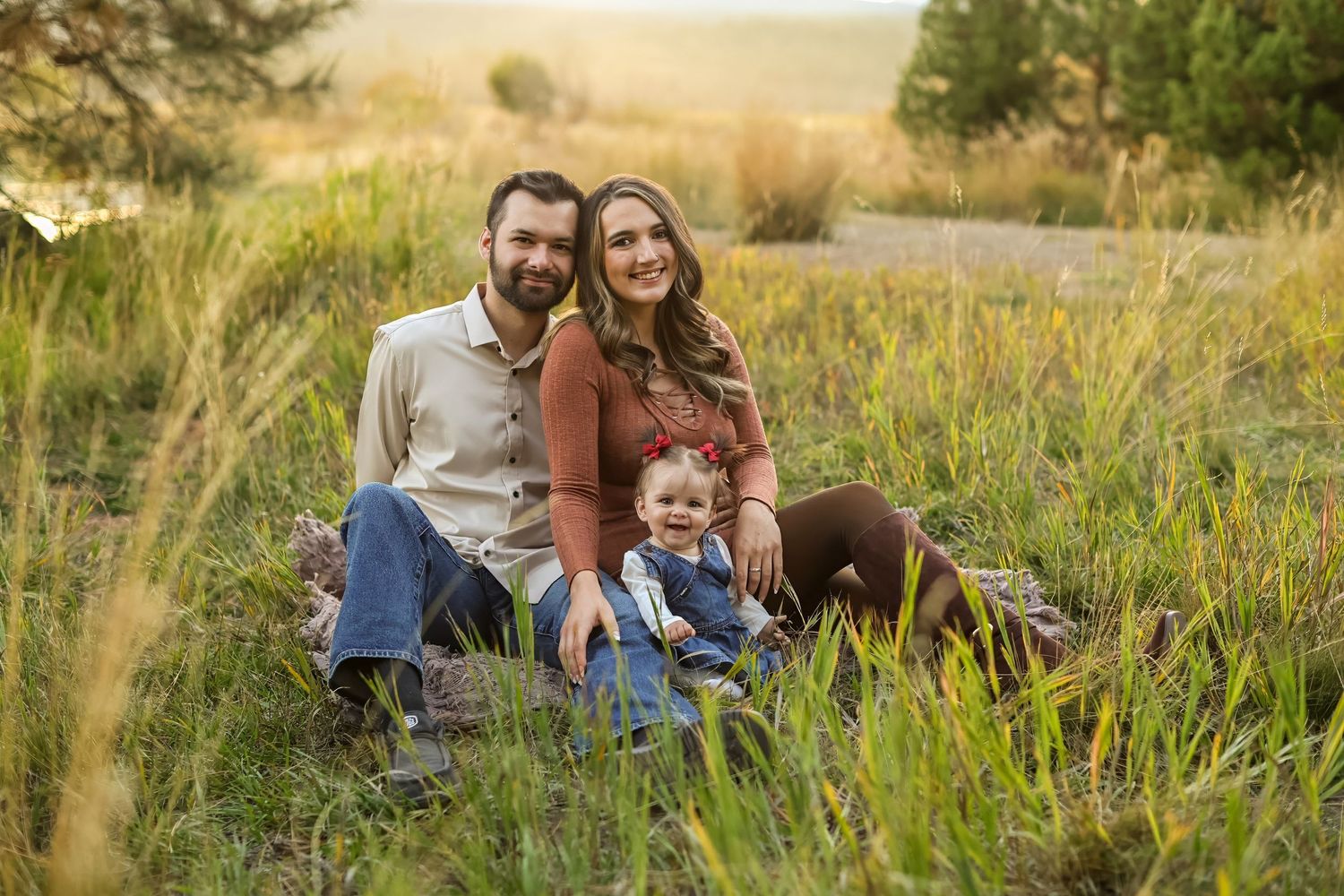A happy family of three sits in a golden meadow, parents smiling with their toddler in a sunlit outdoor portrait.