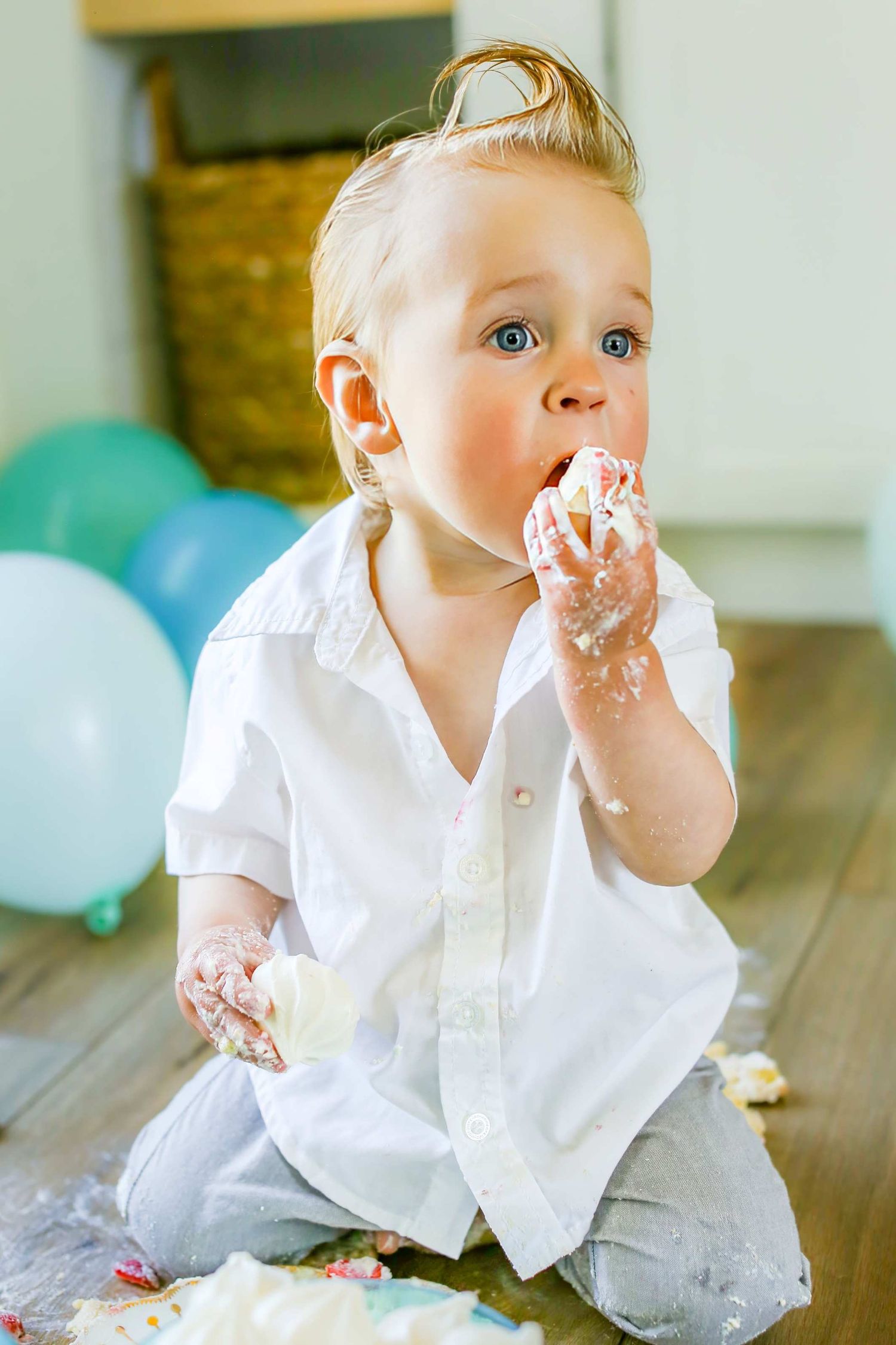 Adorable baby in white shirt eating birthday cake with frosting-covered hands, sitting on floor in Bend Oregon