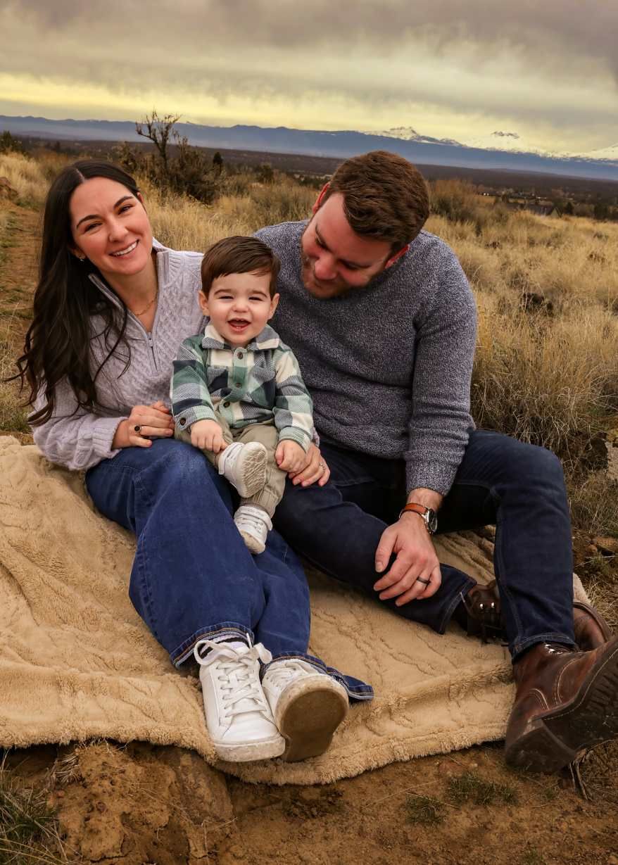 A family sits together on a blanket in a desert landscape with mountains visible in the background at sunset. Redmond Oregon