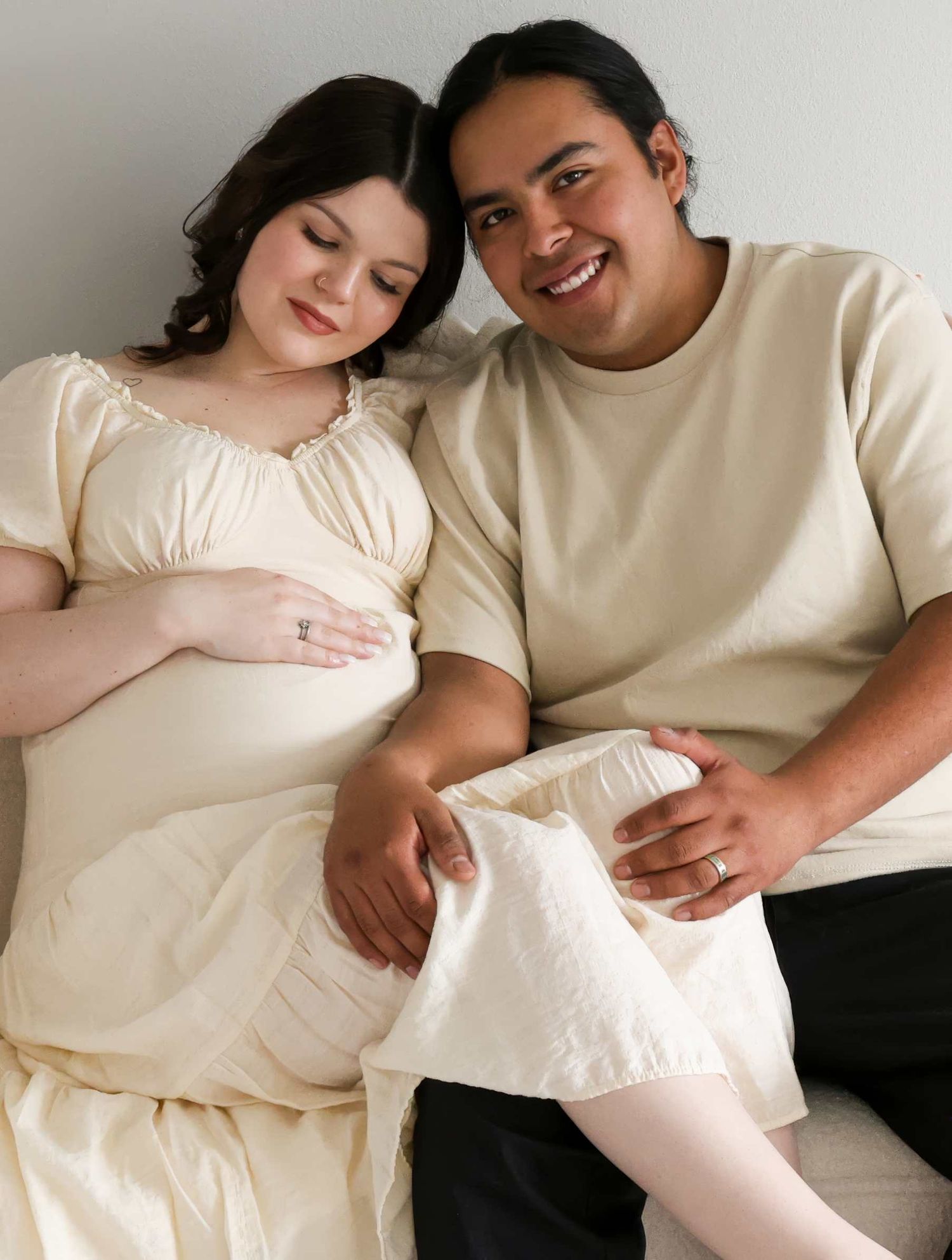 A couple in cream-colored outfits sits together on a couch, sharing intimate moments while expecting a baby. Redmond Oregon Studio