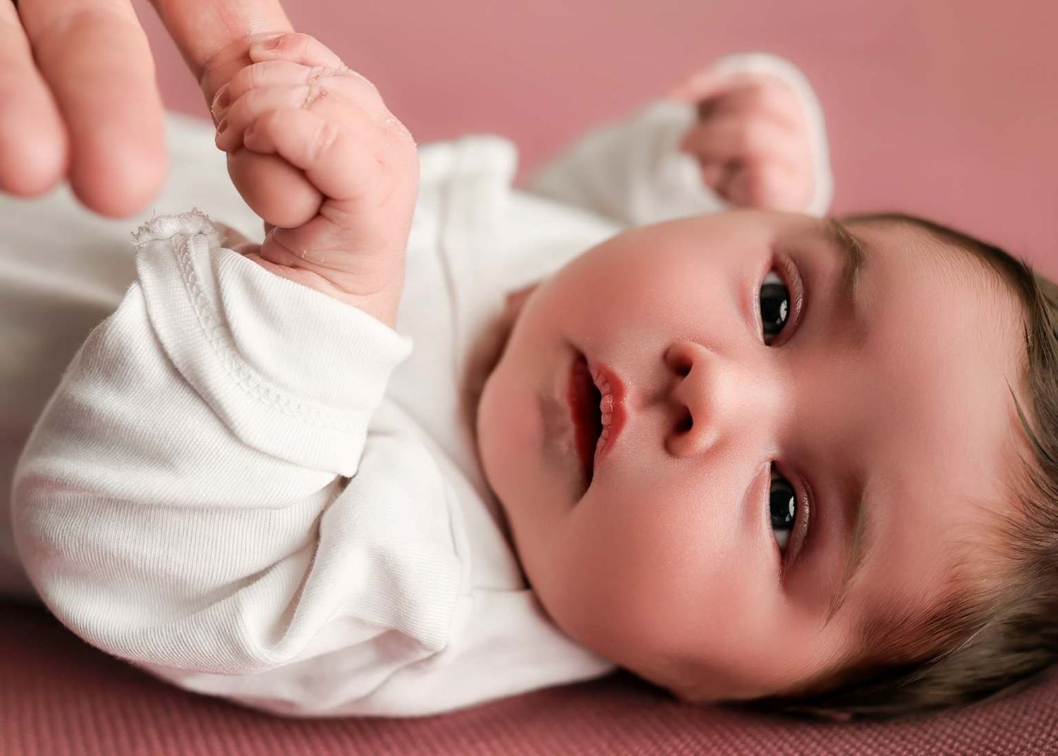 Sweet baby in white onesie lying on pink background in multiple candid poses.