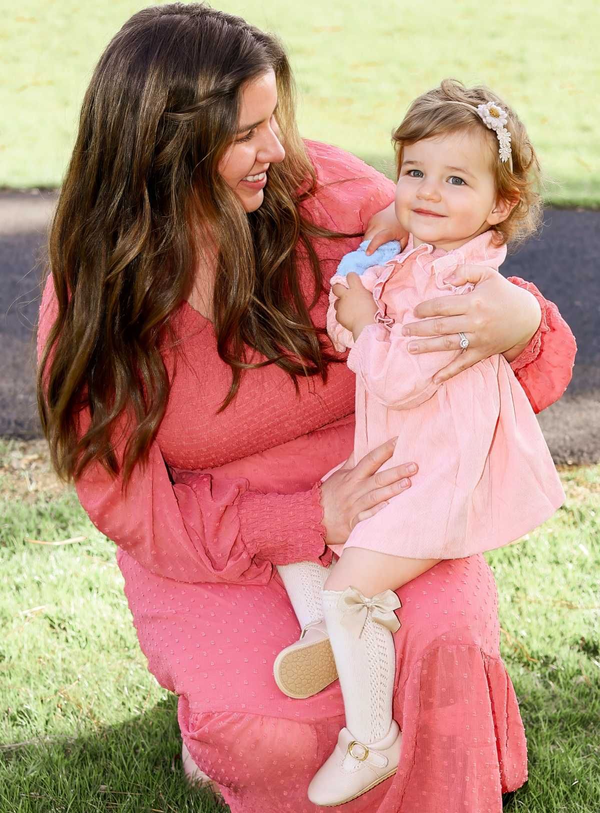 Parent and baby in matching coral pink outfits share joyful moments while playing together outdoors on grass.