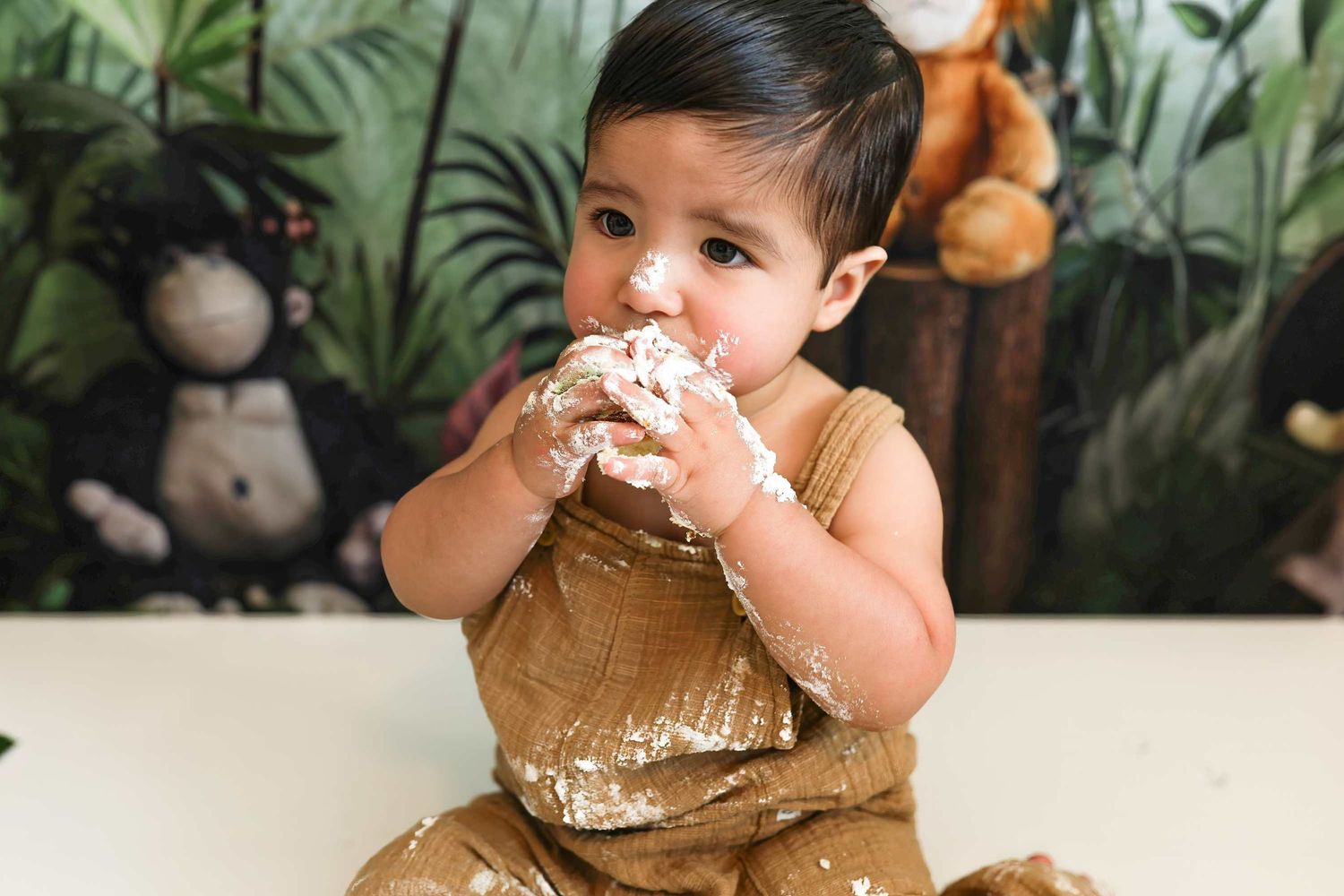 Baby in brown outfit enjoying cake smash photo session with tropical greenery backdrop and orange balloons.