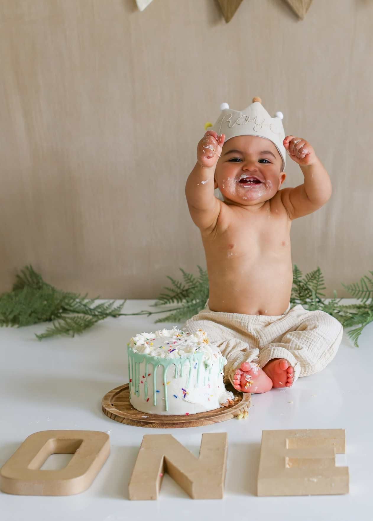 A baby wearing white lace sits among decorative greenery and wooden letters during a cake smash session.