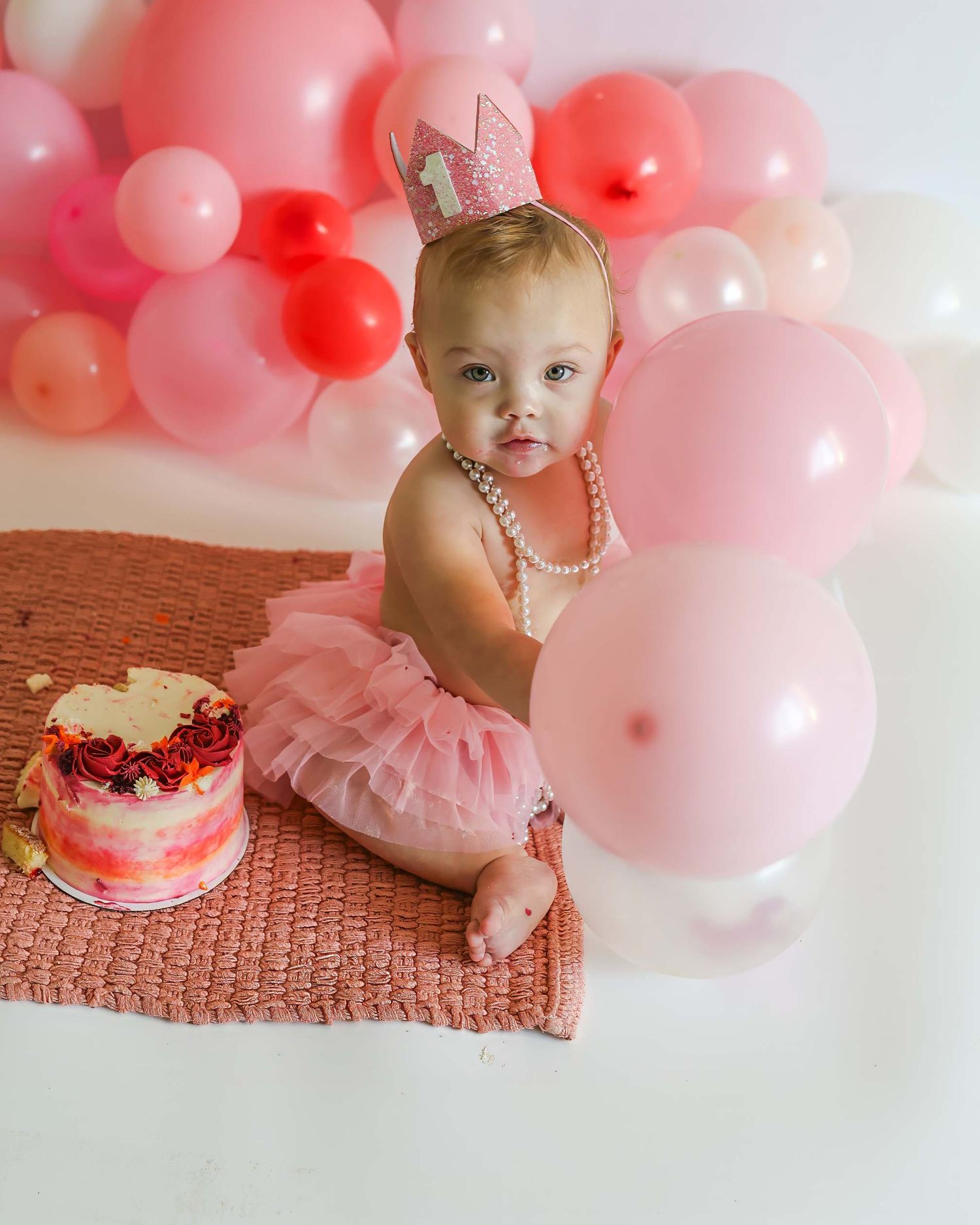 Baby girl in pink tutu and crown sits near smash cake surrounded by pink balloons.