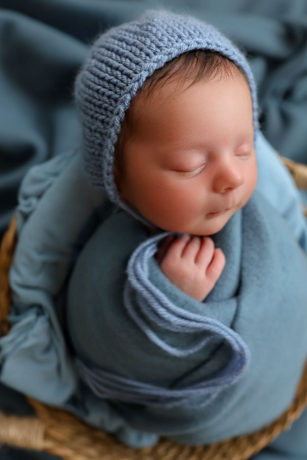 Sleeping newborn baby wrapped in a soft blue knit blanket and matching bonnet, nestled in a wicker basket.