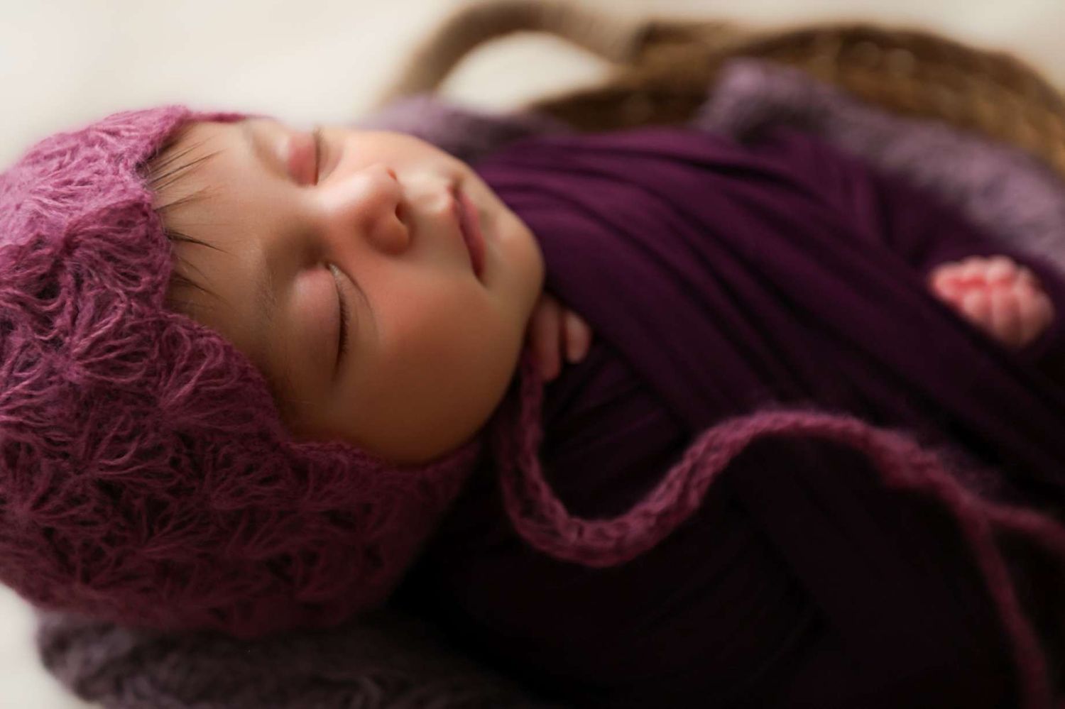 Newborn baby wrapped in purple knit swaddle wearing matching pink bonnet sleeping.