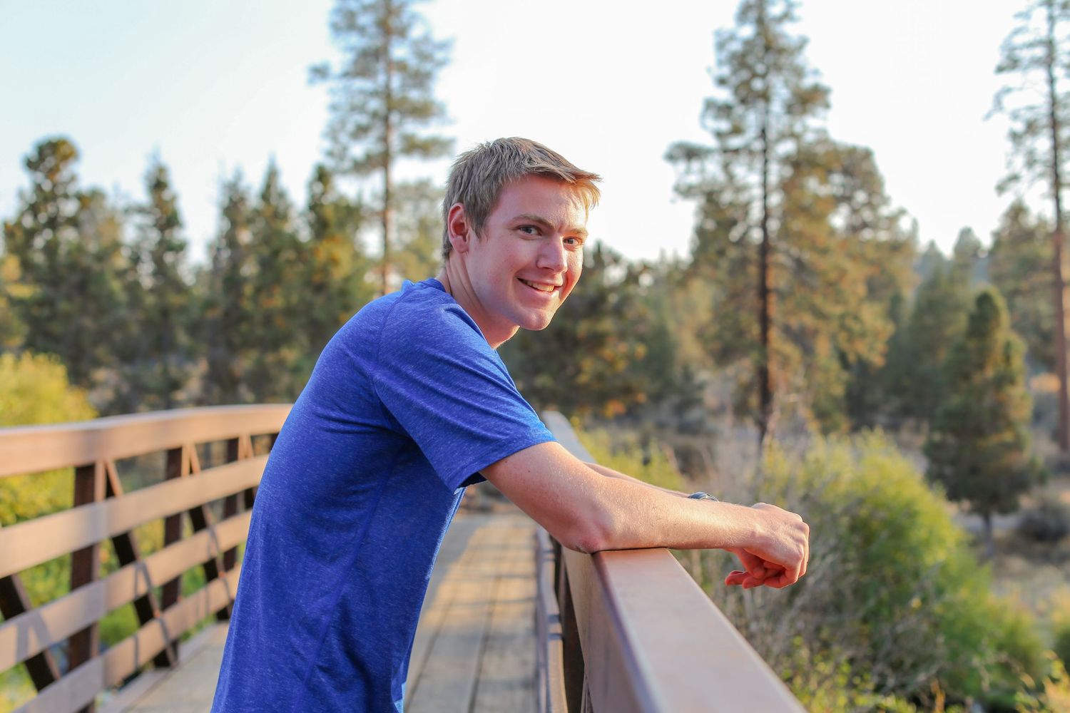 Young man in blue t-shirt smiling on a wooden bridge surrounded by pine trees and natural scenery.