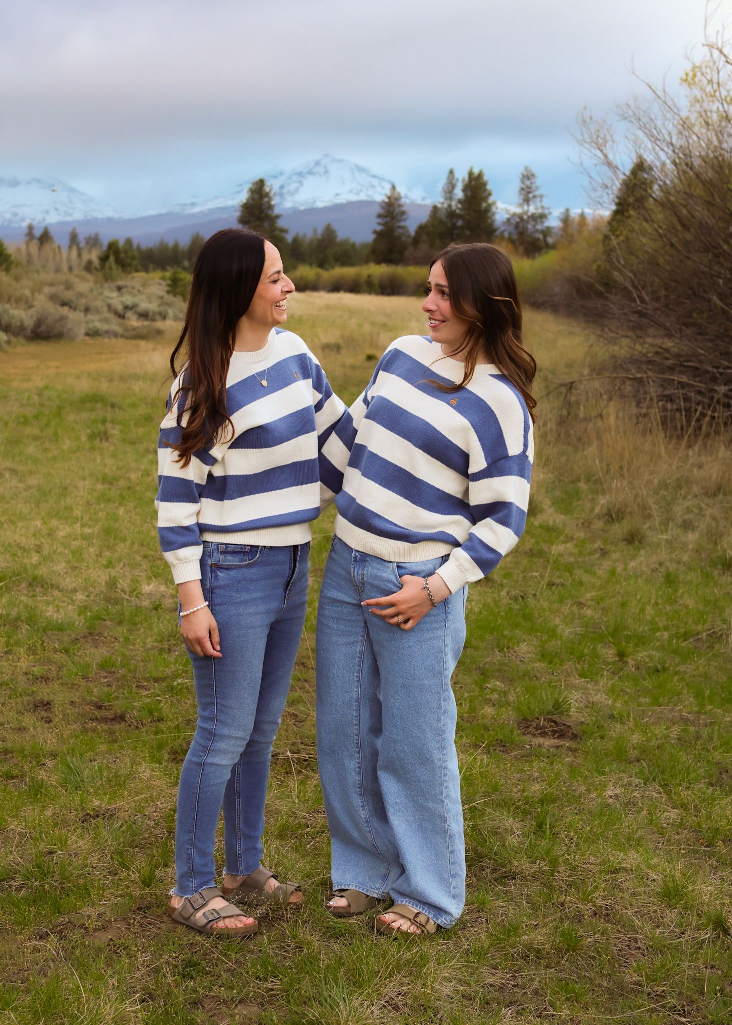Two women in matching blue and white striped sweaters and jeans stand in a mountain meadow, smiling at each other.