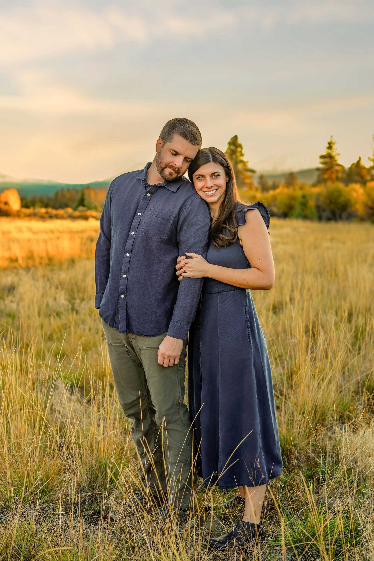 Couple in matching navy outfits embracing in golden meadow at sunset with trees and hills in background.