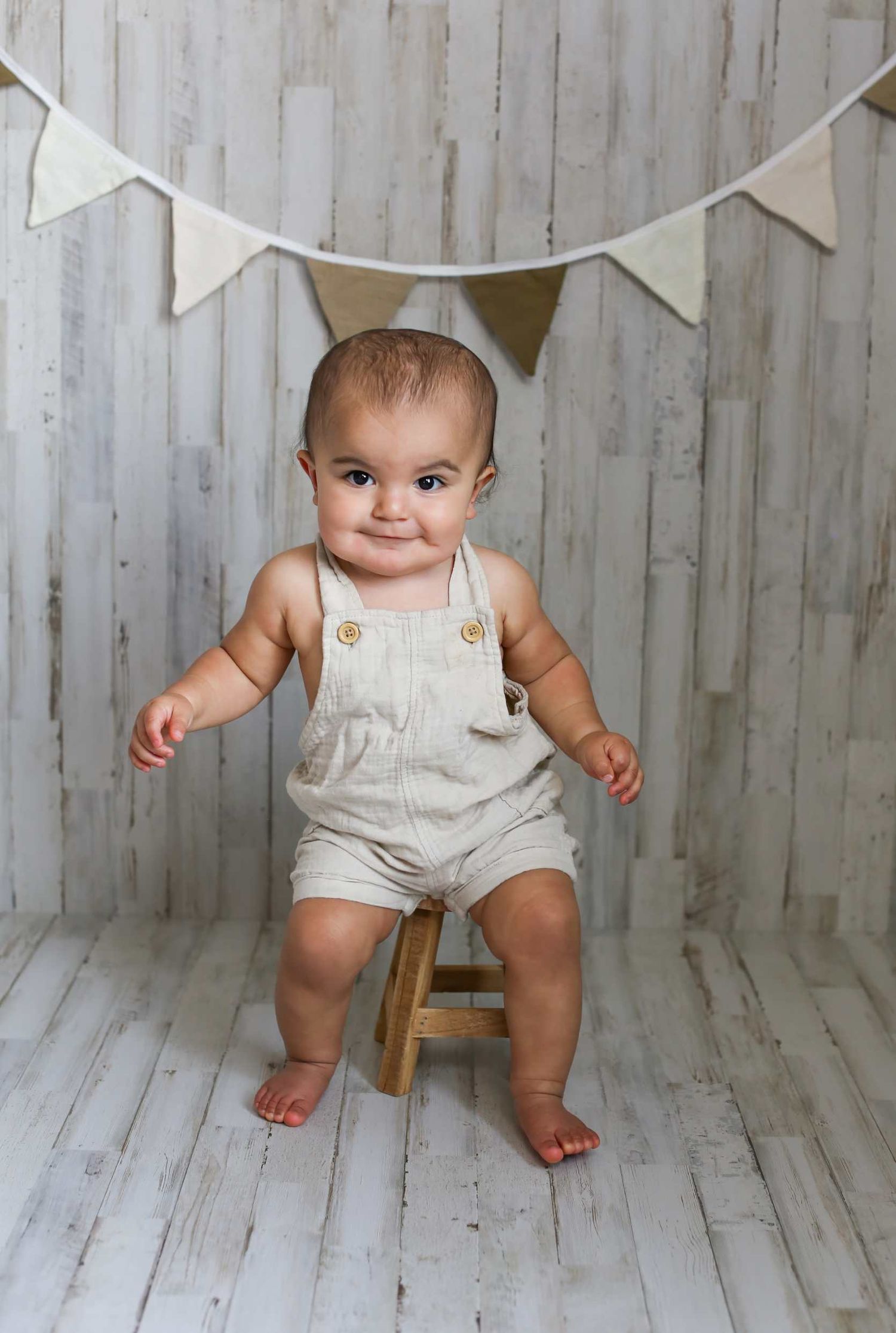 Baby in white overalls sitting on wooden floor with white bunting banner decoration.